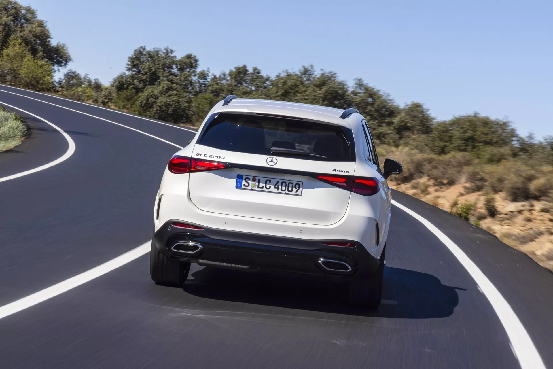 Tylny widok jadącego SUV-a Mercedes-Benz GLC 2022 na zakręconej asfaltowej drodze pod czystym niebem. Pojazd ma jasne metaliczne wykończenie, tylne światła LED, podwójne końcówki wydechu, tylny zderzak w stylu dyfuzora oraz spojler dachowy z wbudowanym światłem hamowania. Widoczne są również wyraźne emblematy i tablica rejestracyjna w stylu europejskim, wskazujące szczegóły modelu i wyposażenia. Wieloramienne felgi i potencjalne funkcje technologiczne, takie jak czujniki parkowania i kamera cofania, dodają luksusowego charakteru.