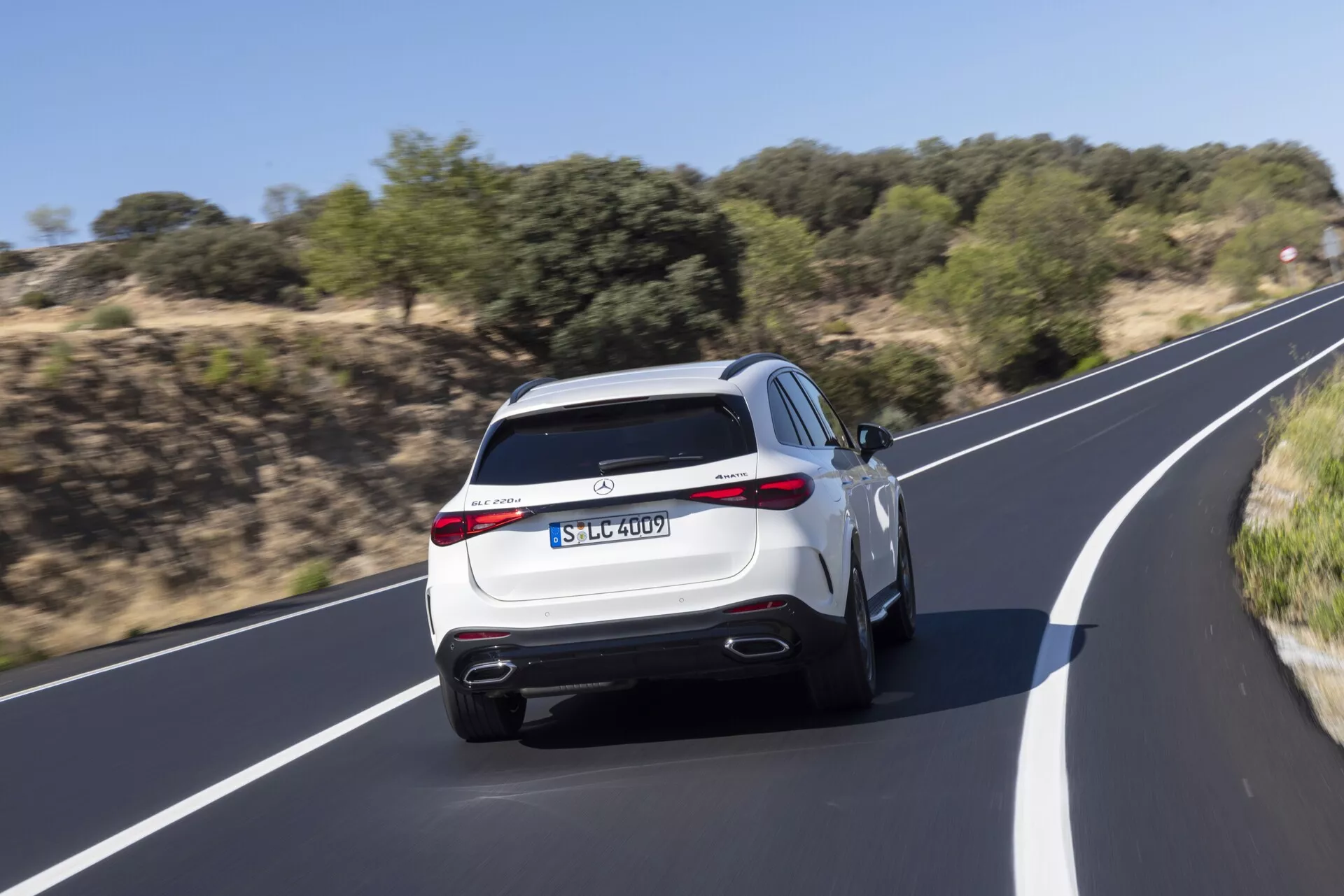 Tylny widok na nowoczesne SUV Mercedes GLC 2022 w ruchu, przedstawiający podwójne wydechy, aerodynamiczny dyfuzor tylny, tylne światła LED, sportowy spojler tylni oraz praktyczne funkcje, takie jak tylny wycieraczek i wysoko umieszczone trzecie światło stop. Jasne malowanie SUV-a, takie jak białe lub srebrne, podkreśla jego ekskluzywny wygląd podczas jazdy po gładkiej asfaltowej drodze otoczonej naturalną roślinnością. Zamazanie ruchu podkreśla dynamiczne osiągi pojazdu.