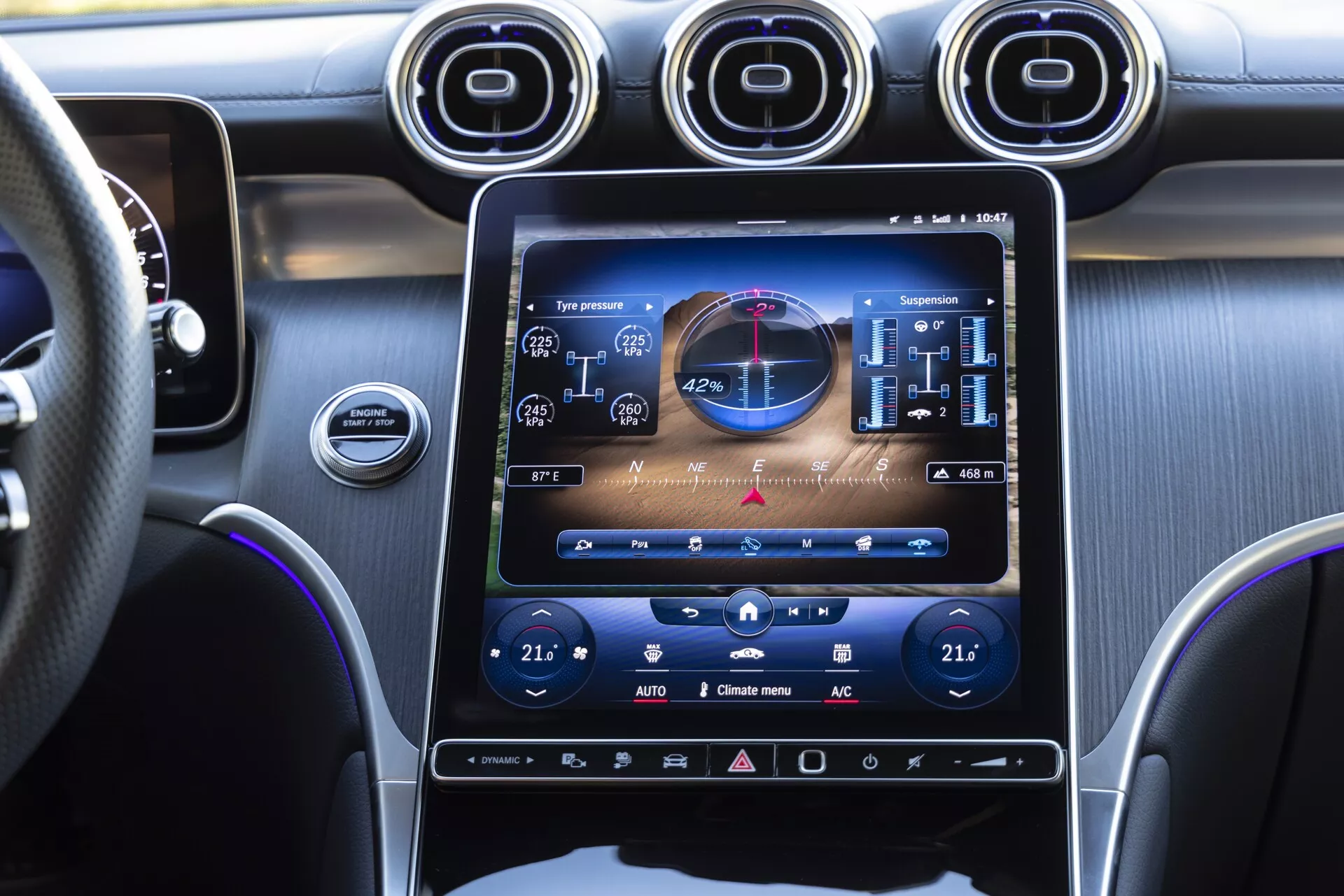 Zbliżenie wnętrza deski rozdzielczej Mercedesa GLC 2022 z perspektywy strony pasażera, podkreślające duży, pionowo zorientowany ekran dotykowy infotainmentu pokazujący funkcje pojazdu, dwustrefową klimatyzację i ustawienia zawieszenia. Widoczne są trzy metalowe, okrągłe kratki wentylacyjne ponad ekranem, kierownica skórzana ze szwami, ciemne tony z wykończeniem ze szczotkowanego metalu oraz dodatkowe dotykowe kontrole pod ekranem, co podkreśla nowoczesny i elegancki design.