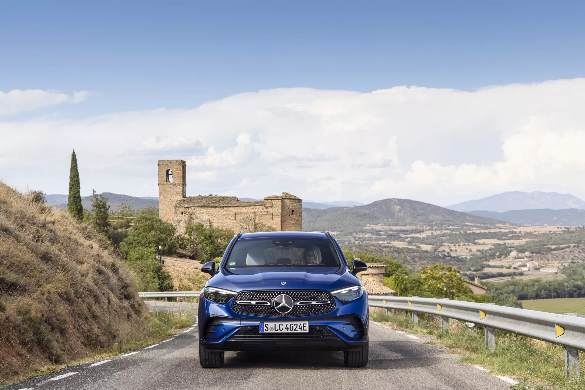 Mercedes-Benz GLC 254 z 2022 roku widziany pod kątem, z widoczną przednią i boczną częścią. Samochód posiada eleganckie reflektory LED, prominentną kratkę i sportowe alufelgi. Zaparkowany na miejskiej ulicy z nowoczesnymi budynkami w tle, w jasnych warunkach dziennych, podkreślających jego metaliczne srebrne wykończenie.