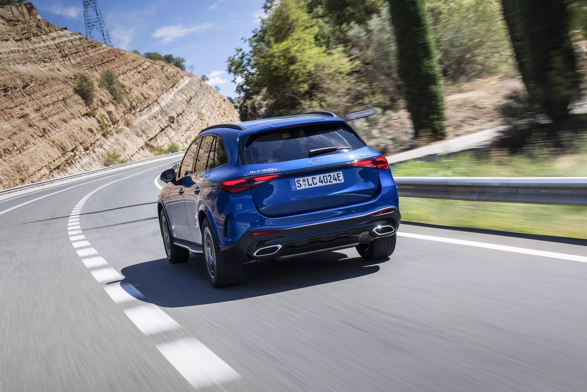 Obraz przedstawia dynamiczny tylni widok z trzech czwartych niebieskiego SUV-a Mercedes GLC 2022 jadącego po asfaltowej drodze zakręcającej w lewo. Uchwycony z lekko podwyższonego kąta, pojazd wyróżnia się chromowanymi końcówkami wydechu, masywnym tylnym zderzakiem z czarnymi akcentami, eleganckimi tylnymi lampami LED oraz stromo pochyloną tylną szybą. Sportowy spojler dachowy, panoramiczny dach i widoczne relingi dachowe dodają atrakcyjności wysokiej klasy. Profil boczny ujawnia wieloszprychowe felgi aluminiowe i opony o niskim profilu, co podkreśla wydajność na drodze. Zaawansowany port ładowania z przodu tylnej lampy i oznaczenie EQ sygnalizują możliwości hybrydowe typu plug-in lub elektryczne, podczas gdy oznaczenie 4MATIC wskazuje na napęd na wszystkie koła. Rozmyte tło z powodu ruchu sugeruje, że pojazd jest w ruchu, co daje wrażenie prędkości i dynamiki.