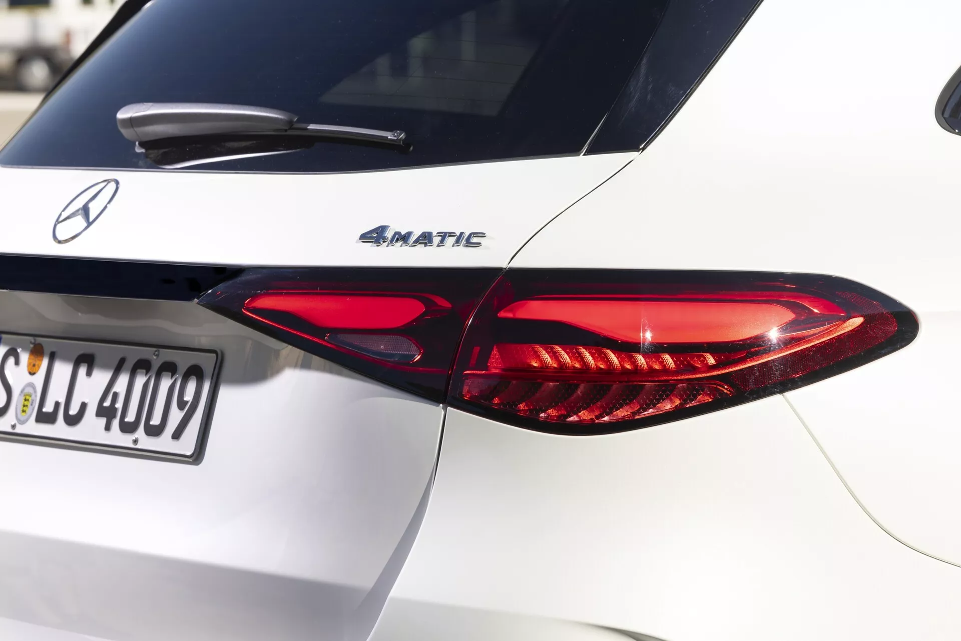Zbliżenie tylnej prawej strony białego Mercedesa-Benz GLC 2022, podkreślające charakterystyczny wydłużony projekt tylnych świateł LED. Tylne światło ma czerwone i przezroczyste sekcje, które mieszczą różne funkcje oświetleniowe. Pokrywa bagażnika pokazuje znaczek modelu '4MATIC', wskazując napęd na wszystkie koła. Naturalne światło słoneczne rzuca cienie na dobrze utrzymane nadwozie samochodu, koncentrując się na tylnym świetle i znaczku z płytką głębią ostrości. Widoczna tablica rejestracyjna zaczyna się od 'SLC'. Wysokiej jakości obraz uchwytujący drobne szczegóły tekstury lakieru i czytelności tekstu, idealny do zilustrowania projektowania samochodów i technologii oświetleniowej.
