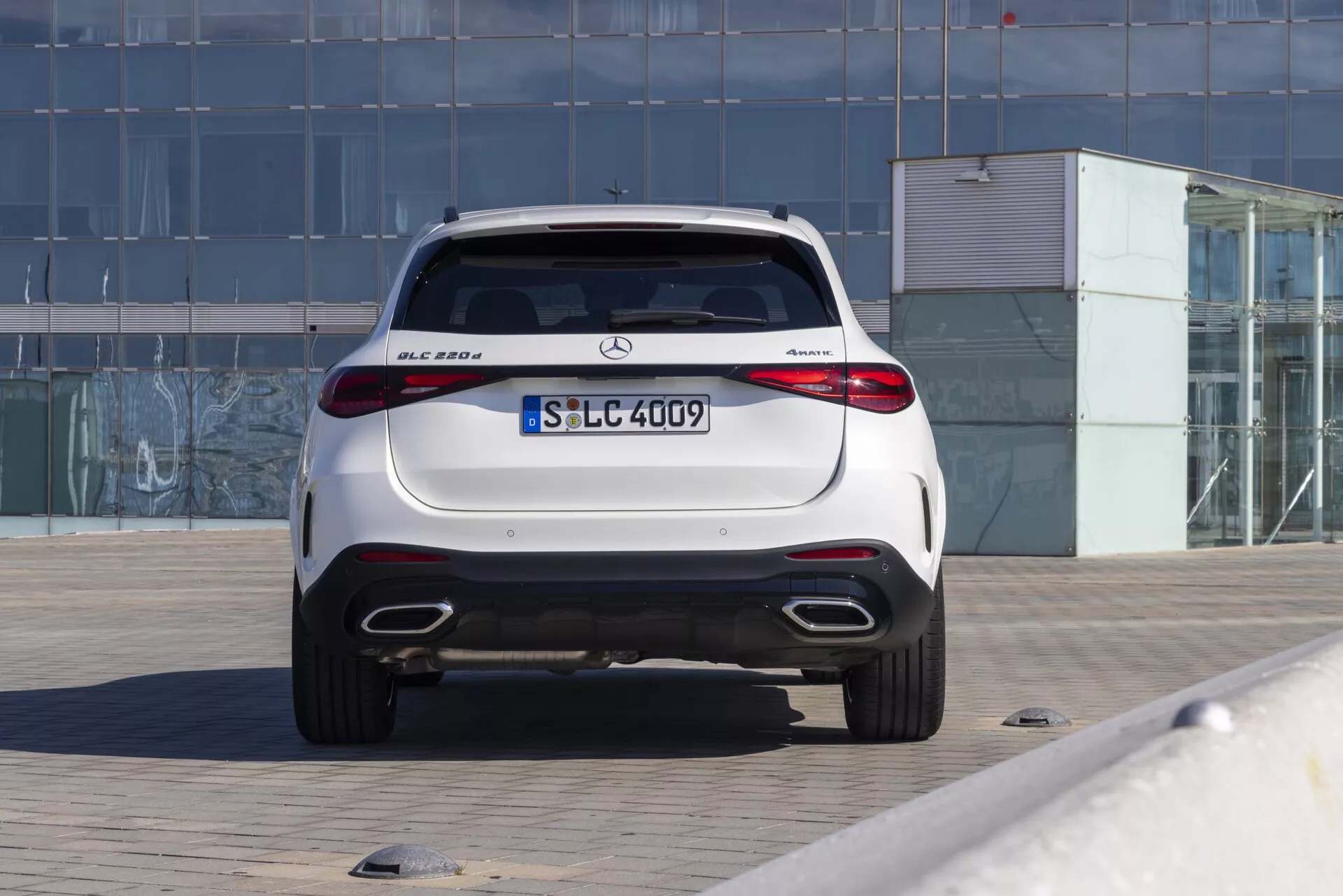 Tylny widok luksusowego kompaktowego SUV-a Mercedes-Benz GLC 300 z 2022 roku z napędem na cztery koła 4MATIC, zaparkowanego na utwardzonym terenie. Fotografia przedstawia aerodynamiczną klapę bagażnika, zintegrowany tylny spojler, poziome światła tylne LED, podwójne rury wydechowe, wbudowane czujniki parkowania, tylny dyfuzor i błyszczące wykończenie. Samochód jest wycentrowany na ziemi, na tle budynku z szklaną elewacją, co wskazuje na miejskie lub komercyjne otoczenie. Dobrze oświetlone zdjęcie podkreśla eleganckie i sportowe cechy designu charakterystyczne dla nowoczesnych pojazdów marki Mercedes-Benz.