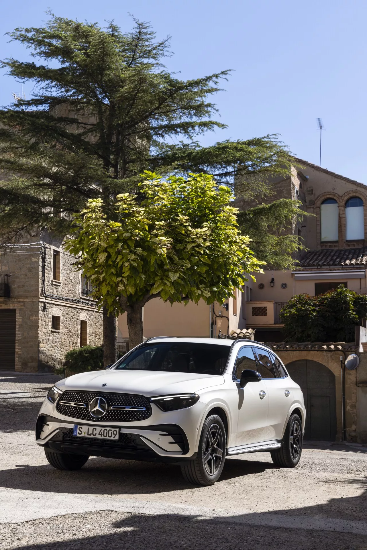 Biały Mercedes-Benz GLC 2022 (254) SUV zaparkowany na brukowanej ulicy w historycznym europejskim mieście, sfotografowany z przedniego trójkątnego kąta. Widoczne funkcje obejmują duże logo Mercedes-Benz na grillu, reflektory LED, aluminiowe felgi, potencjalne kamery/sensory na przednim zderzaku, aerodynamiczny design nadwozia i boczne lusterka mogące mieć dodatkowe kamery lub wskaźniki kierunku. Otoczenie i naturalne światło podkreślają luksusowy wygląd pojazdu.