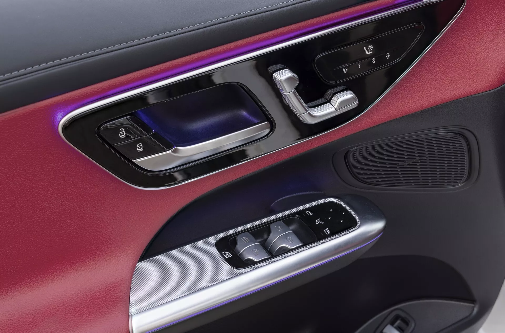 Zbliżenie na panel drzwi kierowcy w Mercedesie GLC (254) z 2022 roku, ukazujący mieszankę materiałów, w tym metalowe wykończenia i matowe czarne powierzchnie. Charakteryzuje się kolorowym oświetleniem ambientowym, ergonomicznie zaprojektowaną klamką drzwi w błyszczącej czerni i metalu, podświetlanymi elektrycznymi przyciskami okien, elektrycznie regulowanymi lusterkami bocznymi oraz zintegrowanym na dole głośnikiem wysokiej jakości, podkreślając luksusowy i technologicznie zaawansowany wystrój wnętrza samochodu.