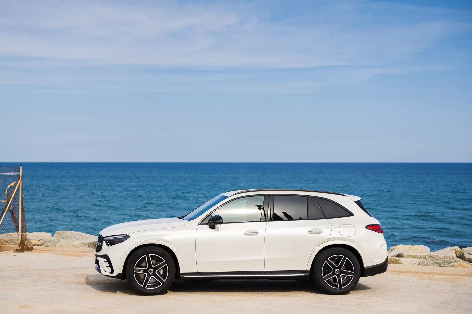 Obraz białego luksusowego samochodu typu crossover Mercedes GLC 2022 (254) zaparkowanego na utwardzonym terenie. Widok z boku podkreśla profil pasażerskiego boku pojazdu z widocznymi przednimi i tylnymi kołami wieloramiennymi. W tle widać morze i czyste niebo. Elegancki design SUV obejmuje lusterka boczne, klamki drzwi i prawdopodobnie szyberdach. Cienie pod pojazdem wskazują na słoneczne warunki. Lekko przyciemniane szyby dodają luksusowego wyglądu.