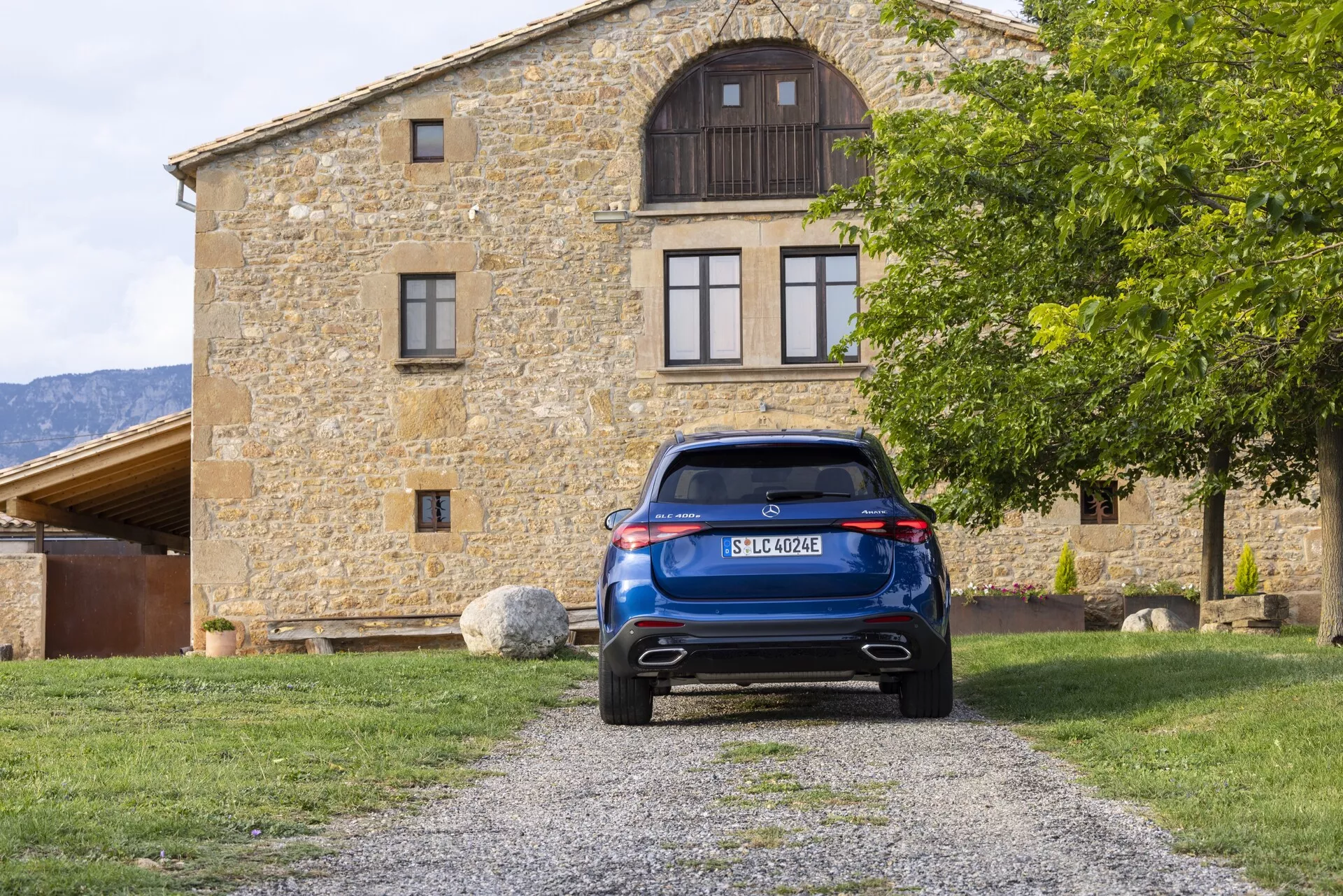 Widok z tyłu niebieskiego Mercedesa GLC 2022 (254) zaparkowanego na żwirowej ścieżce, wycentrowany w kompozycji z budynkiem w tle. Obraz podkreśla klapę tylną, tylnią szybę z wycieraczką, nowoczesne tylne światła LED, tylny zderzak z czujnikami parkowania lub odblaskami oraz podwójny układ wydechowy. Funkcje obejmują wysokie światło stop, wyśrodkowaną tablicę rejestracyjną, element dyfuzora i spojler dachowy, ukazując tym samym nowoczesną technologię i design motoryzacyjny.
