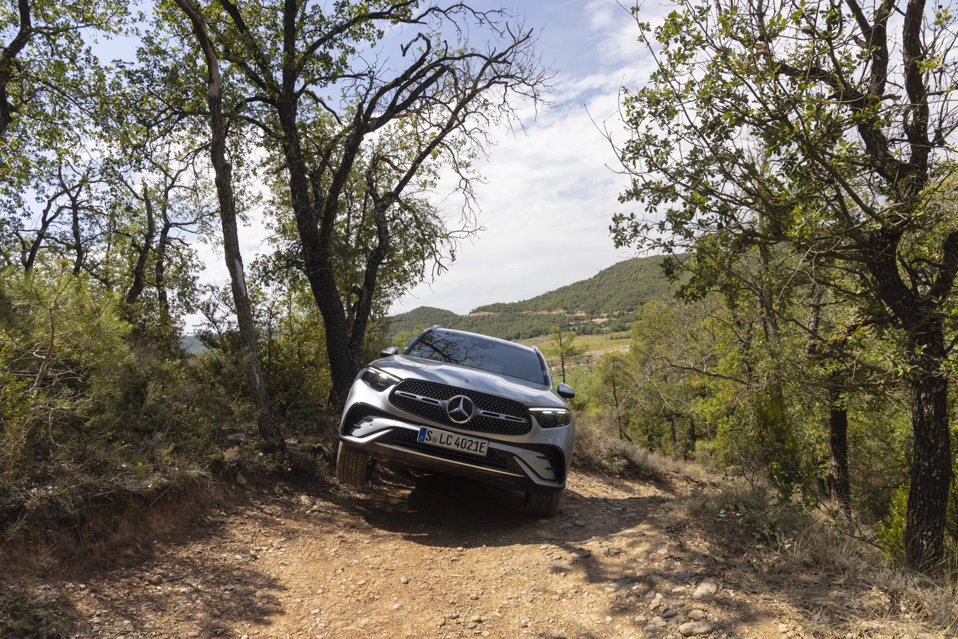 Kolorowe zdjęcie SUV Mercedes-Benz GLC 2022 w naturalnym otoczeniu, uchwycone z niskiego kąta przedniego, podkreślając jego surowy wygląd. Zdjęcie koncentruje się na przedniej fasadzie samochodu, grillu i nowoczesnych reflektorach LED. Pojazd znajduje się na ścieżce gruntowej, otoczony zielenią, co podkreśla jego terenowe możliwości i elegancki design. Przednie czujniki i kamery sugerują zaawansowane funkcje bezpieczeństwa.