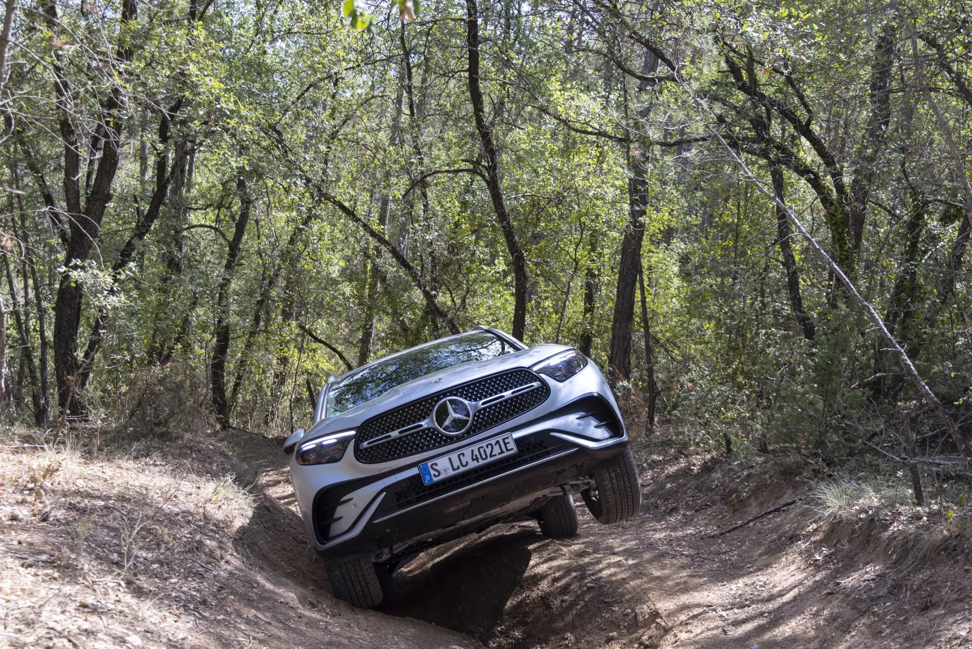 SUV Mercedes-Benz GLC 2022 na trudnym, leśnym szlaku terenowym widziany z trójkątnego kąta przedniego, pokazujący pełną kratkę, emblemat, reflektory, część maski i przedni zderzak z osłoną; strona pasażera bardziej widoczna z przechyłem na nierównym terenie, otoczony drzewami i krzakami ukazującymi możliwości terenowe SUV.