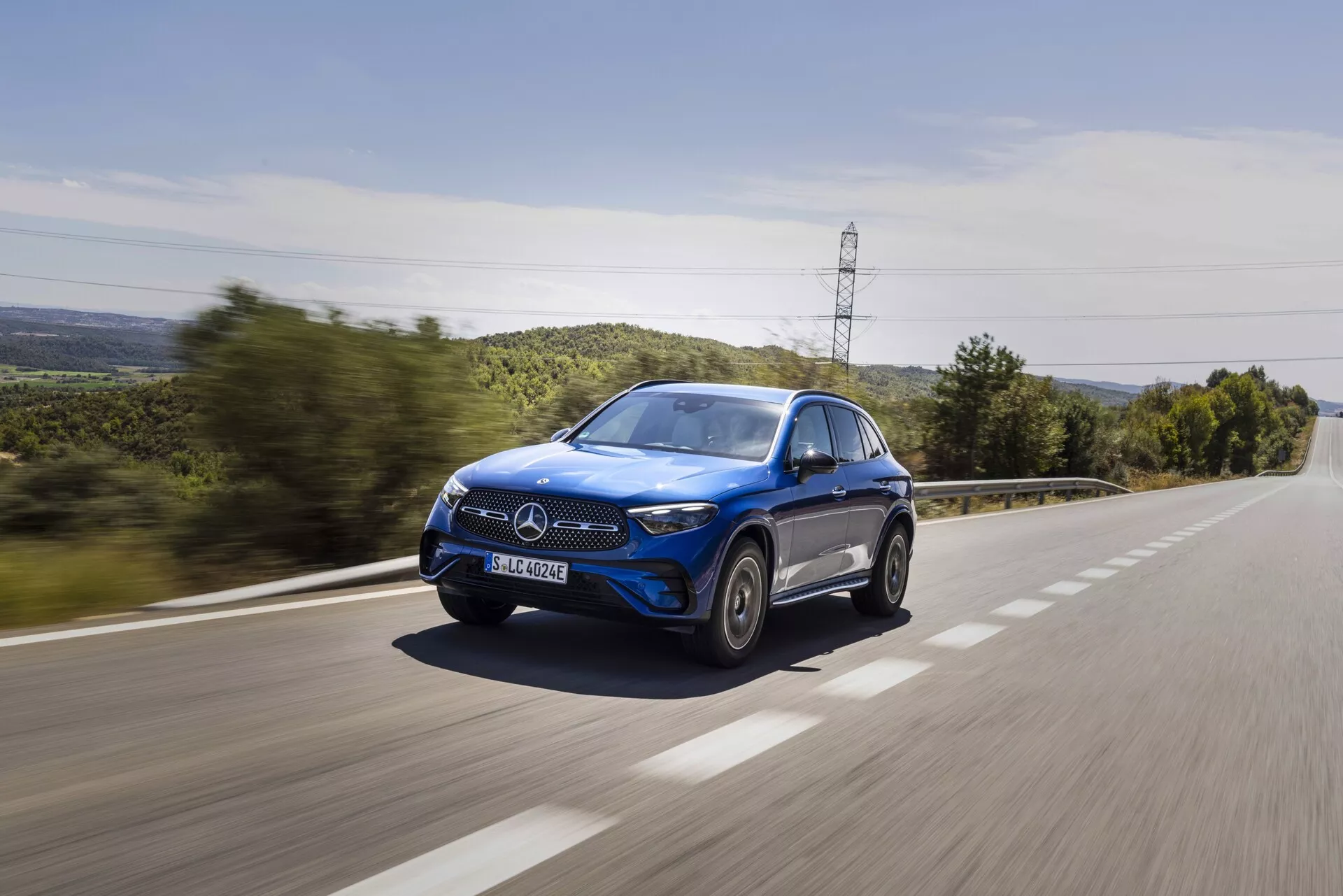 Niebieski SUV Mercedes-Benz GLC 2022 jadący z dużą prędkością po wiejskiej drodze, uchwycony z trzy-czwartego przedniego kąta, ukazujący przedni grill, maskę, reflektory LED, lusterko strony kierowcy, koła i okna, z efektem rozmycia ruchu na powierzchni drogi i zielenią w tle, podkreślający dynamiczny i luksusowy design auta.