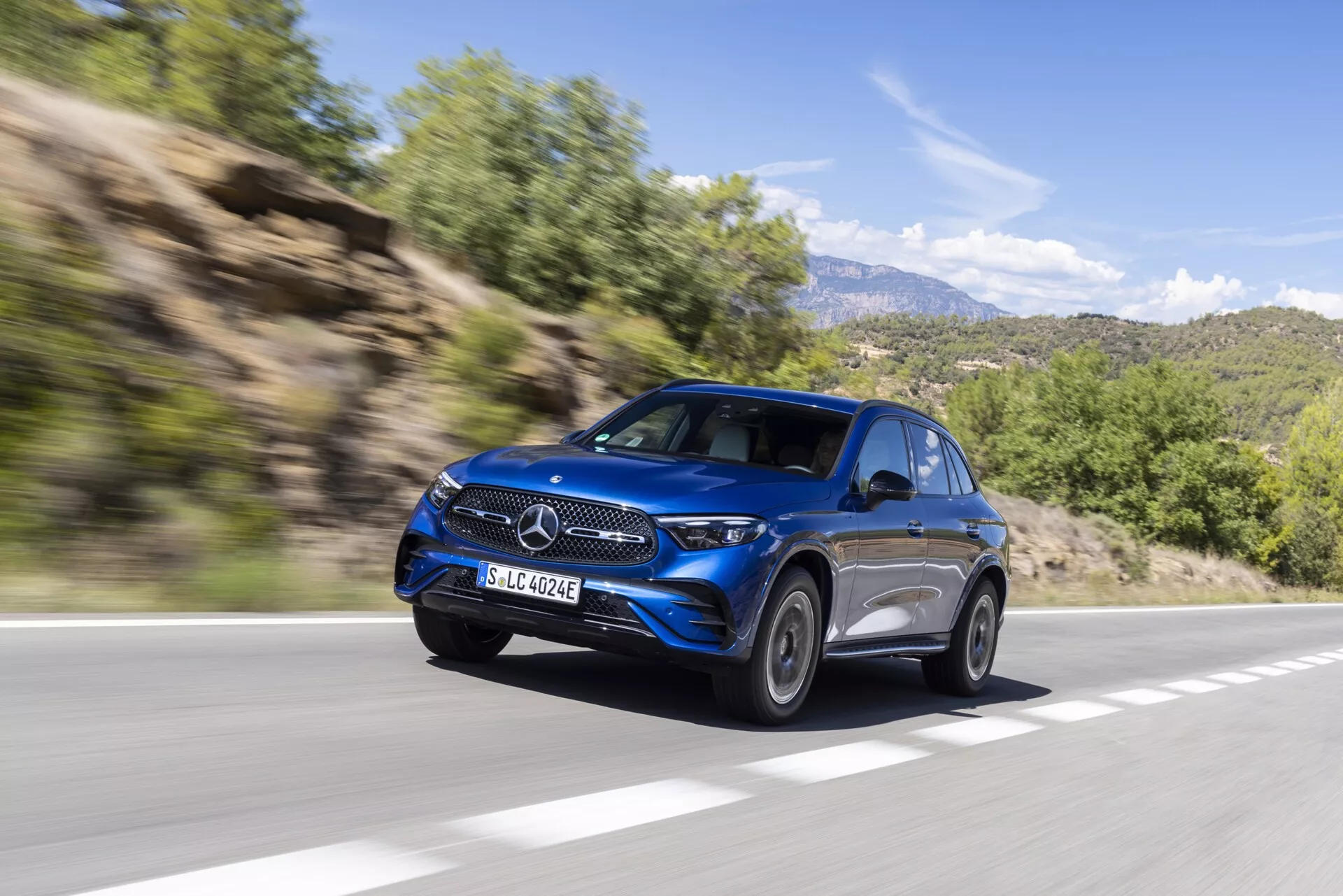 Dynamiczne ujęcie niebieskiego SUV-a Mercedes GLC 2022 (254) w ruchu, widzianego z kąta przedniego lewego. Obraz ukazuje jego eleganckie reflektory, wyrazistą kratkę z centralnym emblematem, luksusowe felgi aluminiowe, aerodynamiczne linie nadwozia i zaawansowane lusterka boczne. Pojazd jedzie po asfaltowej drodze w górskim terenie wiejskim, na tle rozmytego przez ruch liści i skalistych wzgórz, dodając wrażenia prędkości i nowoczesnego luksusu.