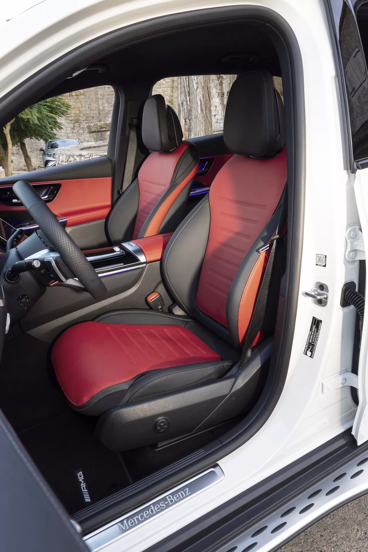 Widok wnętrza Mercedes-Benz GLC 2022, ukazujący dwubarwne sportowe przednie fotele z lśniącą skórą, zaawansowany panel sterowania siedzeniami na otwartych drzwiach kierowcy oraz częściowo widoczną zaawansowaną wielofunkcyjną kierownicę i elegancką deskę rozdzielczą z możliwością personalizacji oświetlenia ambientowego.