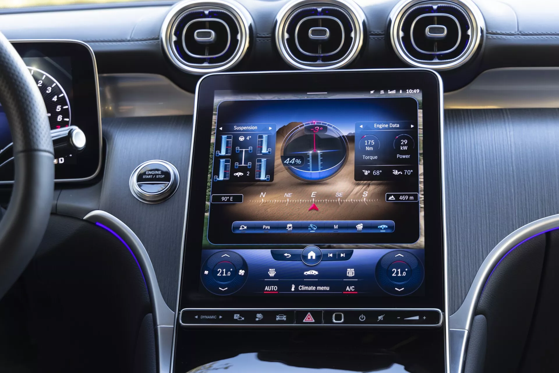 Widok na poziomie oczu z przedniego fotela pasażera we wnętrzu Mercedesa GLC 2022, ukazujący konsolę centralną z dużym pionowym ekranem dotykowym systemu informacyjno-rozrywkowego wyświetlającym GPS, audio i elementy sterowania klimatyzacją. Trzy metalowe okrągłe otwory wentylacyjne powyżej ekranu, zarys panelu wskaźników i kierownica po lewej stronie, z fioletowym oświetleniem akcentowym podkreślającym konsolę i deskę rozdzielczą.