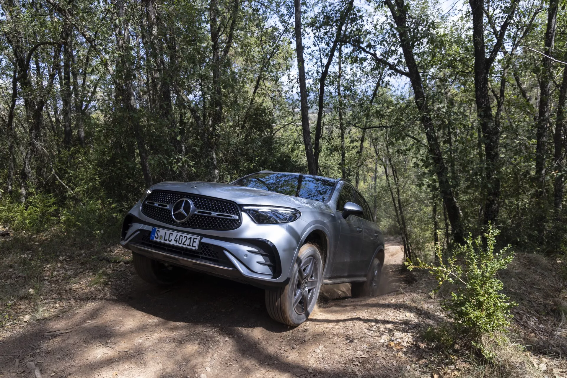 SUV Mercedes GLC 2022 na drodze gruntowej otoczonej zalesionym terenem, uchwycony pod kątem trzech czwartych z przodu, podkreślający jego wyrazisty grill z emblematem marki, reflektory LED, aluminiowe felgi i wysokie prześwity, podkreślające jego zdolności terenowe w naturalnym otoczeniu drzew i suchego terenu.