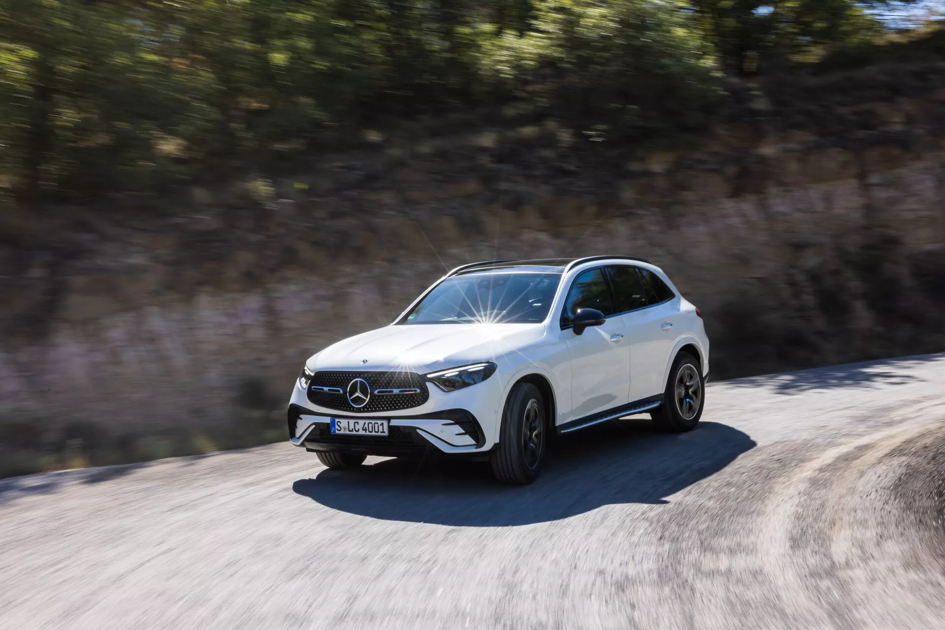 Dynamiczne ujęcie białego SUV-a Mercedes-Benz GLC 2022 w ruchu, uchwycone za dnia z trzech czwartych przedniej strony. Uwagi dotyczące wzornictwa : przedni grill z widocznym emblematem, charakterystyczne reflektory LED, przedni zderzak z widocznymi wlotami powietrza, rzeźbiona maska z unoszącymi się liniami oraz boczny profil do tylnych drzwi. Koła przedstawione w ruchu z lekkim rozmyciem, co sugeruje prędkość. Lusterka boczne są zaprojektowane aerodynamicznie. Otoczenie wydaje się rozmyte z powodu ruchu pojazdu, podkreślając dynamiczny i szybki charakter samochodu.