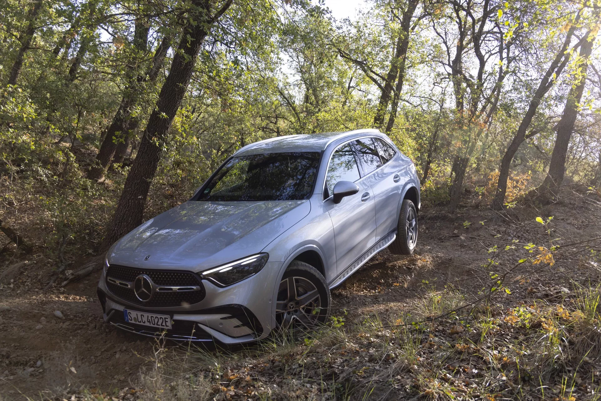 Obraz srebrnego SUV-a Mercedes GLC 2022, ustawionego na zewnątrz w lesistym terenie, pokazując jego zdolności terenowe. Pojazd jest ustawiony pod kątem w lewo, zapewniając widok z trzech czwartych przodu i bocznego profilu, w tym przedniego grilla, ostrych reflektorów LED, konturowanego przedniego zderzaka, maski, felg aluminiowych z bieżnikiem terenowym i płynnych linii. Naturalne światło słoneczne prześwituje przez drzewa, podkreślając nowoczesny, aerodynamiczny design i sugerując zaawansowane funkcje wspomagania kierowcy umieszczone za emblematem grilla.