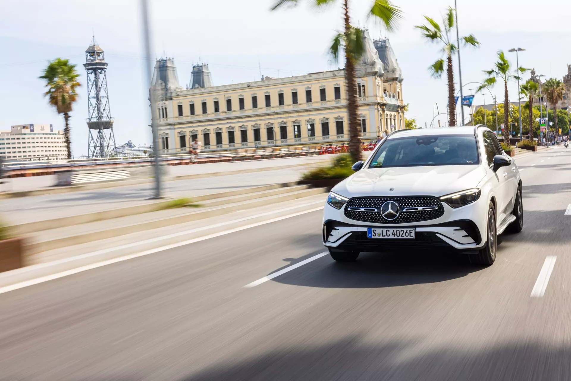Mercedes-Benz GLC 2022 (254) luksusowy sedan elektryczny w białym kolorze z czarnym dachem, uchwycony w ruchu w dziennym otoczeniu miejskim z palmami i zabytkowymi budynkami w tle, z zastosowaniem techniki panoramowania, aby utrzymać ostrość pojazdu na tle rozmytego tła. Z charakterystycznym emblem na grille’u Mercedes-Benz, czujnikami wspomagania kierowcy, cienkimi światłami do jazdy dziennej, eleganckimi reflektorami LED i zintegrowanymi kierunkowskazami w lusterkach.