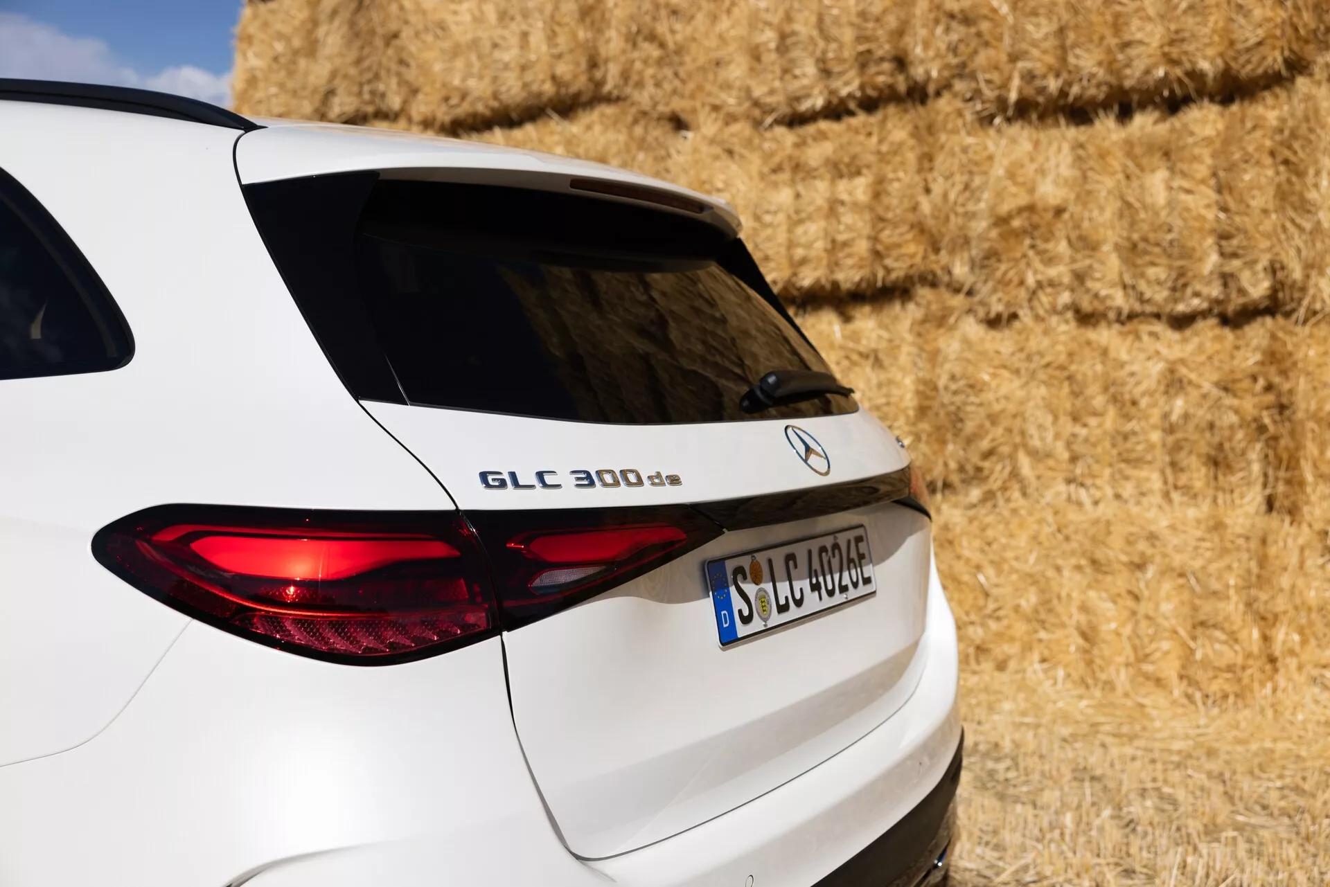 Zbliżenie tylnego widoku trzy czwarte białego SUV-a Mercedes-Benz GLC 300 d, ukazujące prawą stronę tyłu, w tym klapę tylnią, podświetlone tylne światło i część tylnych okien. Pojazd jest zaparkowany w wiejskim otoczeniu z balotami siana w tle. Widoczne cechy obejmują nazwę modelu 'GLC 300 d', logo Mercedes-Benz oraz tablicę rejestracyjną w formacie europejskim, podkreślając eleganckie linie projektu i nowoczesne oświetlenie pojazdu.