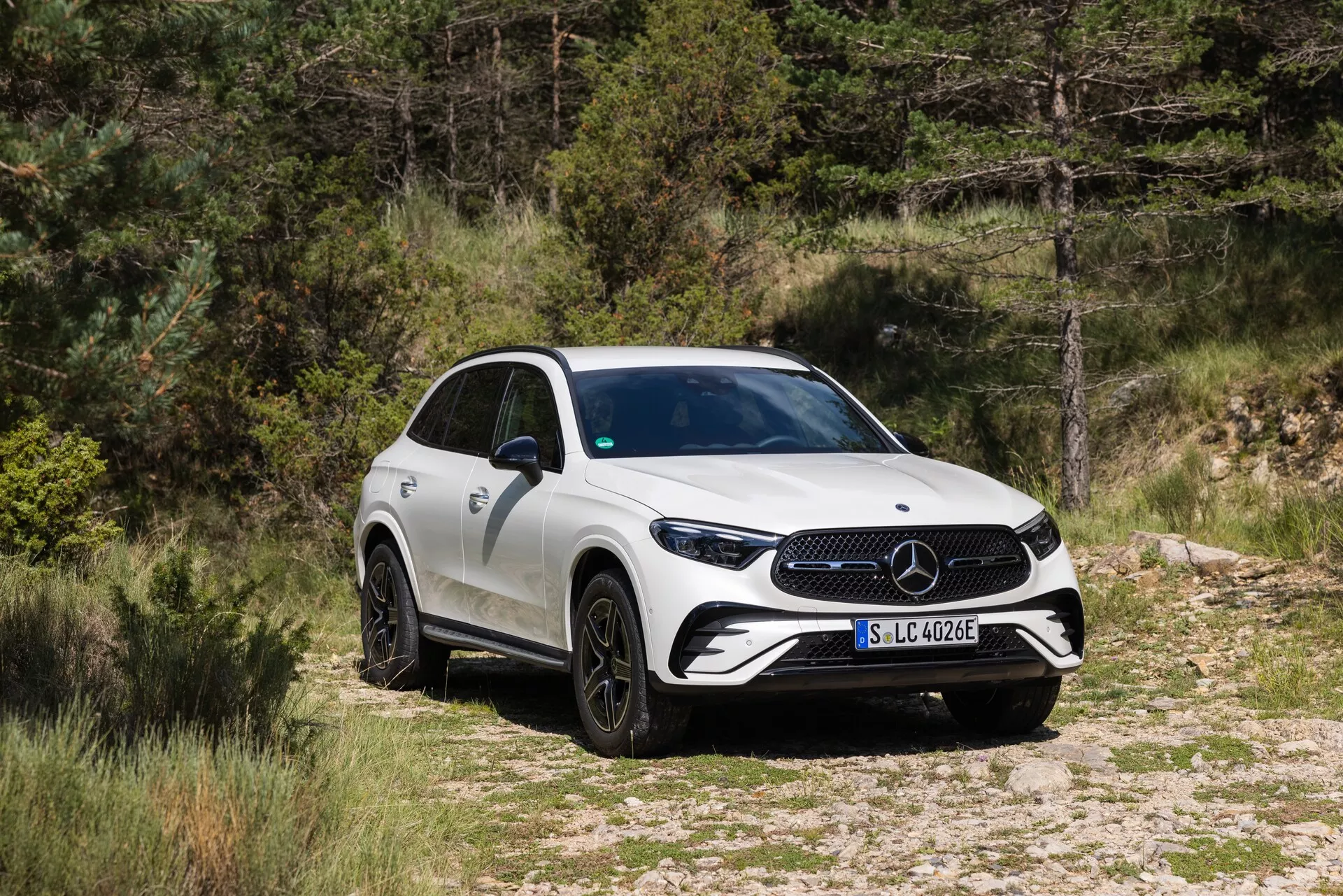 Obraz SUV-a Mercedes-Benz GLC 2022 w naturalnym otoczeniu, usytuowanego na drodze gruntowej lub żwirowej z roślinnością w tle. Pojazd uchwycony z widoku trzech czwartych z przodu, prezentujący przedni grille z dużym znakiem, ostre reflektory LED i część bocznej karoserii. SUV posiada wieloszprychowe, czarne felgi aluminiowe, lusterka boczne z wbudowanymi kierunkowskazami i wznoszącą się linię pasa, prowadzącą do sportowego, zwężającego się tyłu. Karoseria jest w jednolitym białym kolorze, a obraz sugeruje, że ten model jest częścią elektrycznej linii Mercedes-Benz, co wskazuje odznaka 'EQ' na przednim błotniku.