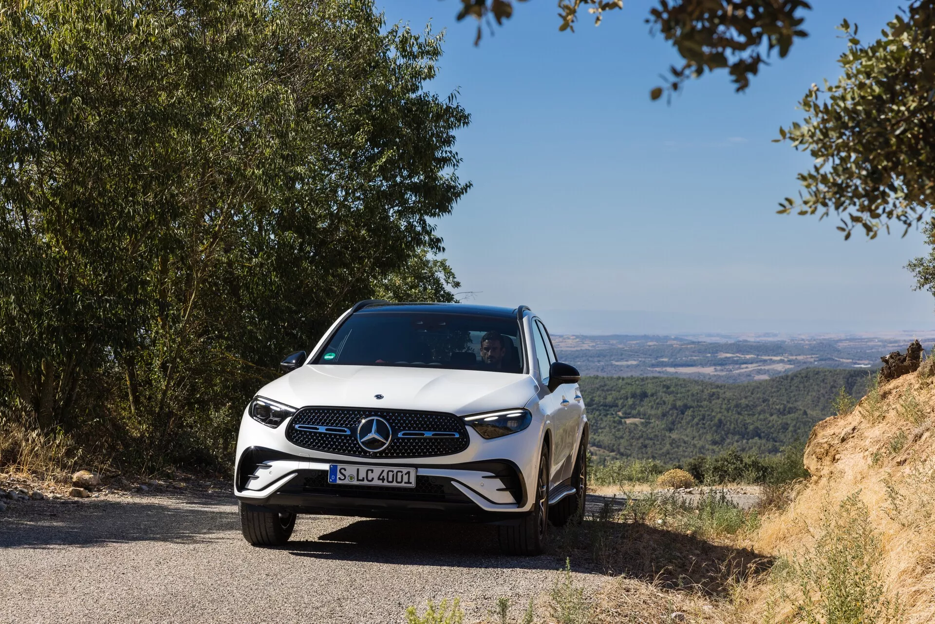 Nowoczesny Mercedes-Benz GLC 2022 jest ustawiony na wąskiej drodze z naturalnym, pagórkowatym tłem, które daje widok na dolinę lub równiny. Widoczny jest przedni znak i design grilla pojazdu, uchwycony z niskiej, frontalnej perspektywy, nieco z lewej strony, podkreślając przedni grill, reflektory i część maski. Reflektory LED, charakterystyczna kratka z dużym emblematem i sportowy design zderzaka podkreślają zaawansowane i eleganckie cechy samochodu. Zdjęcie w ciągu dnia z naturalnym oświetleniem przywołuje spokój, pokazując zaawansowane systemy wspomagania kierowcy implikowane przez emblemat. Elegancki, aerodynamiczny profil jest zaakcentowany liniami maski, które łączą się z rzeźbionymi bokami, uchwyconymi z odległości około 3-5 metrów od kamery.