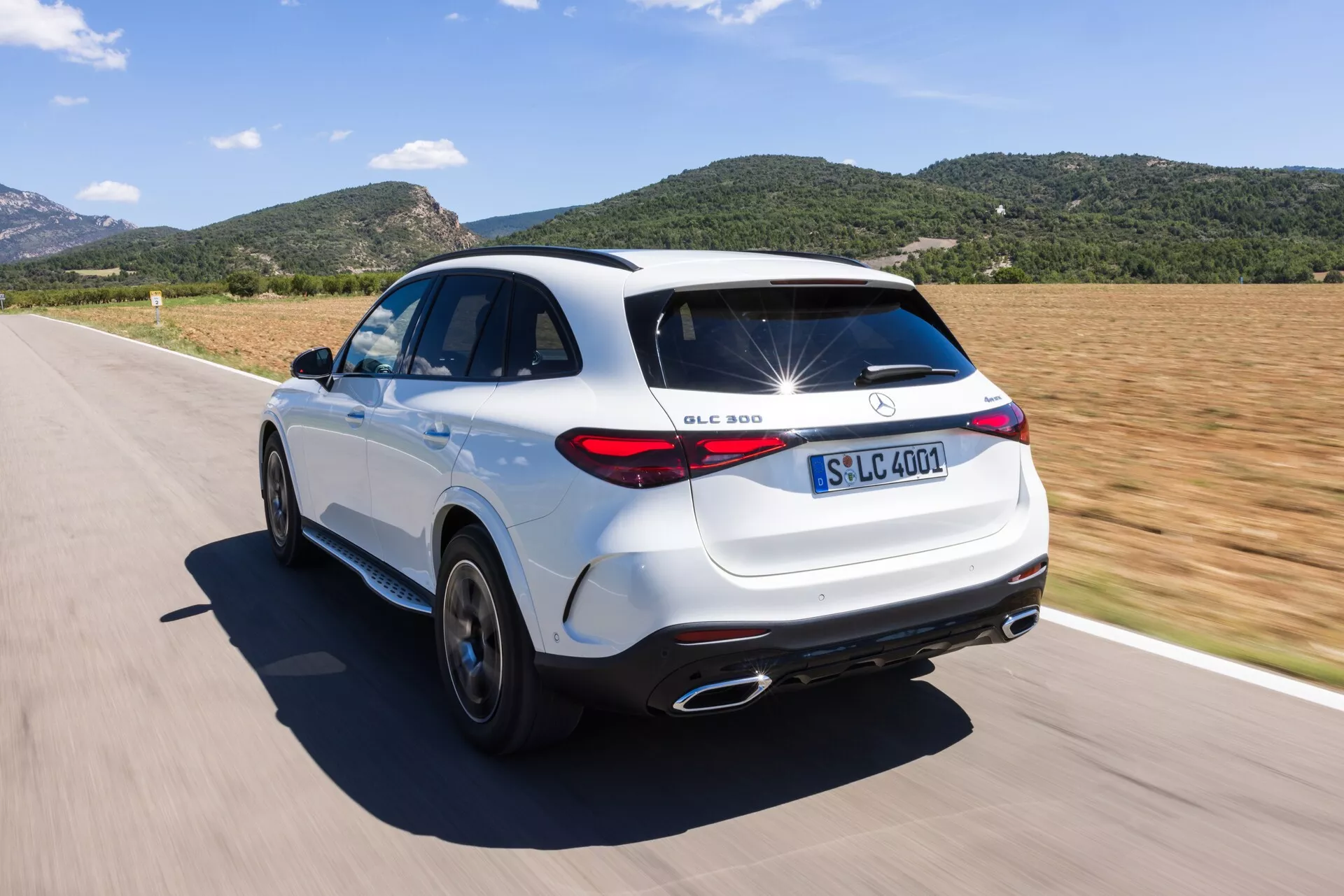 Widok z wysoka, z tyłu pod kątem trzech czwartych białego SUV-a Mercedes-Benz GLC 300 na drodze. Zdjęcie pokazuje cechy projektowe, takie jak charakterystyczne znaki, tylne światła LED, chromowane końcówki wydechów, pięcioramienne felgi aluminiowe i charakterystyczny dachowy spoiler. SUV jest w ruchu w słoneczny dzień, podkreślając jego dynamiczne możliwości i sportową estetykę.