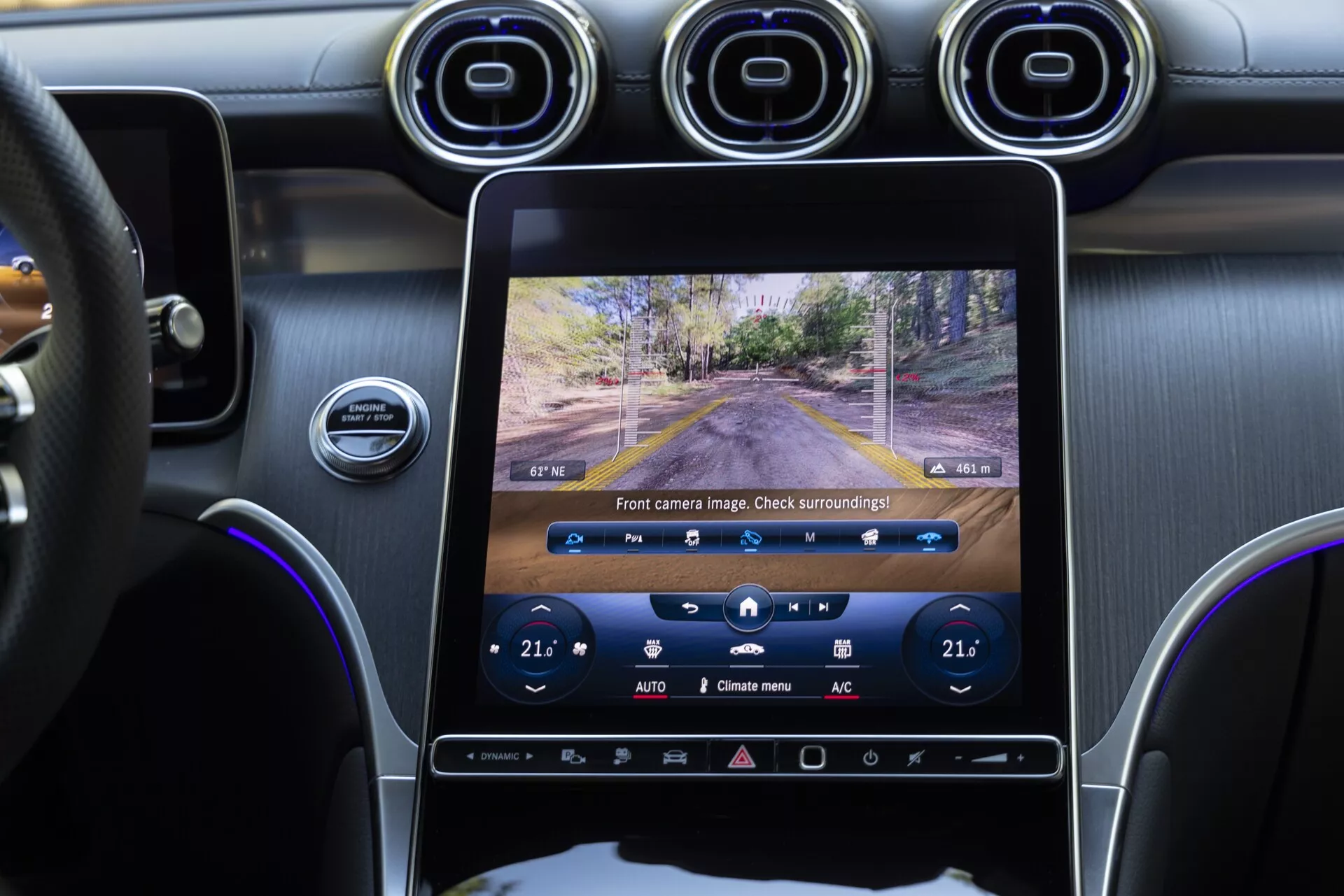Widok wnętrza Mercedesa GLC 2022 skupiający się na konsoli środkowej i systemie multimedialnym, zrobiony z perspektywy tylnego pasażera. Duży pionowy ekran dotykowy pokazuje na żywo obraz kamery przedniej, z widocznym niżej sterowaniem klimatyzacją ustawionym na 21°C. Widoczne są również deska rozdzielcza, kierownica i część drzwi kierowcy, pokazując elegancką kombinację interfejsów cyfrowych i fizycznych przycisków, okrągłe metalowe nawiewy oraz kierownicę obszytą skórą.
