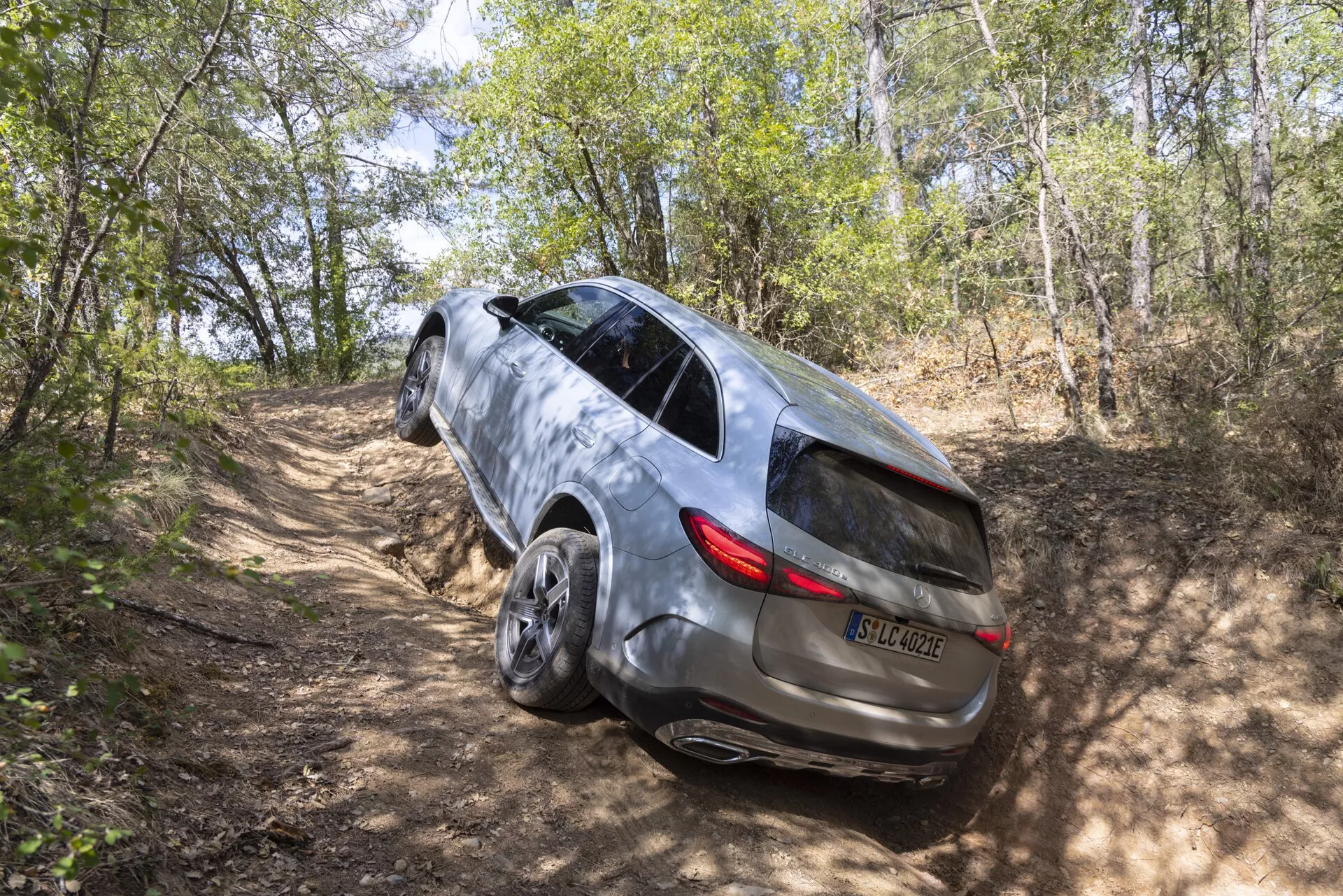 Widok z wysokiego kąta trzy czwarte tyłu na SUV Mercedes GLC 2022 na drodze gruntowej w lesistej okolicy, prezentujący jego zdolność do jazdy na wszystkie koła, elegancki design z panoramicznym dachem, tylne światła LED, metaliczną powłokę lakierniczą, przyciemniane boczne okna z wbudowanymi światłami kierunkowskazów, wpuszczane klamki drzwi i dużymi felgami ze stopów.