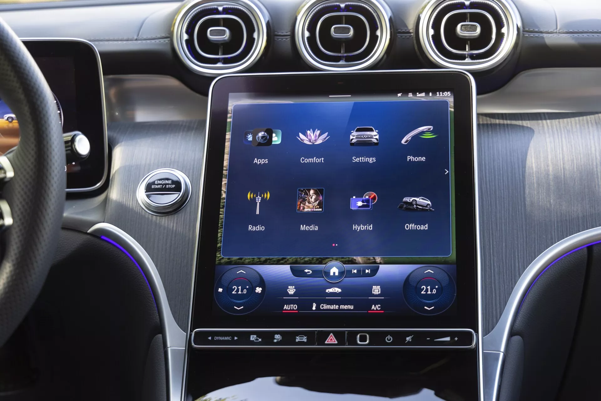 Centralna konsola Mercedesa-Benz GLC 2022 widziana z miejsca kierowcy, skupiająca się na dużym pionowym ekranie dotykowym systemu informacyjno-rozrywkowego, który pokazuje opcje takie jak 'Aplikacje,' 'Komfort,' 'Ustawienia,' wraz z dwustrefową kontrolą klimatu, metalowymi otworami wentylacyjnymi, przyciskiem start/stop oraz premium mieszanką szczotkowanych metalowych i błyszczących czarnych wykończeń.