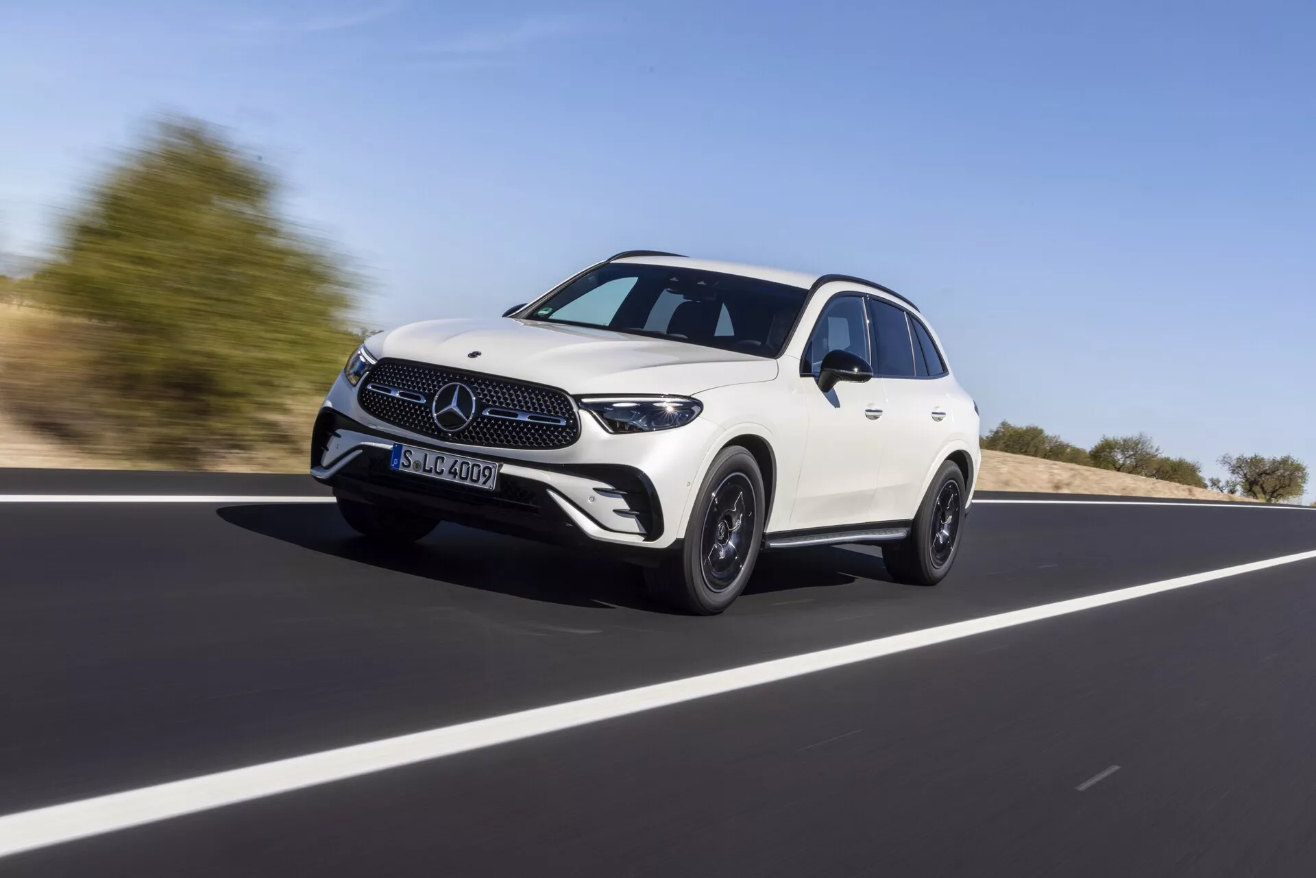 Mercedes GLC 2022 (254) biały czterodrzwiowy crossover SUV w ruchu na otwartej drodze, uchwycony z niskiego kąta podkreślającego jego wysokość i sylwetkę. Pojazd porusza się po przekątnej od lewej do prawej, a uwaga skupiona jest na przedniej kracie, reflektorach, LEDowych światłach do jazdy dziennej i dolnym przednim zderzaku. Profil boczny ukazuje wyprofilowaną linię dachu, linie projektowe, poszerzone nadkola i felgi aluminiowe, łącząc luksus i sportowy charakter. Rozmyte tło sugeruje prędkość, pod jasnym dziennym niebem.