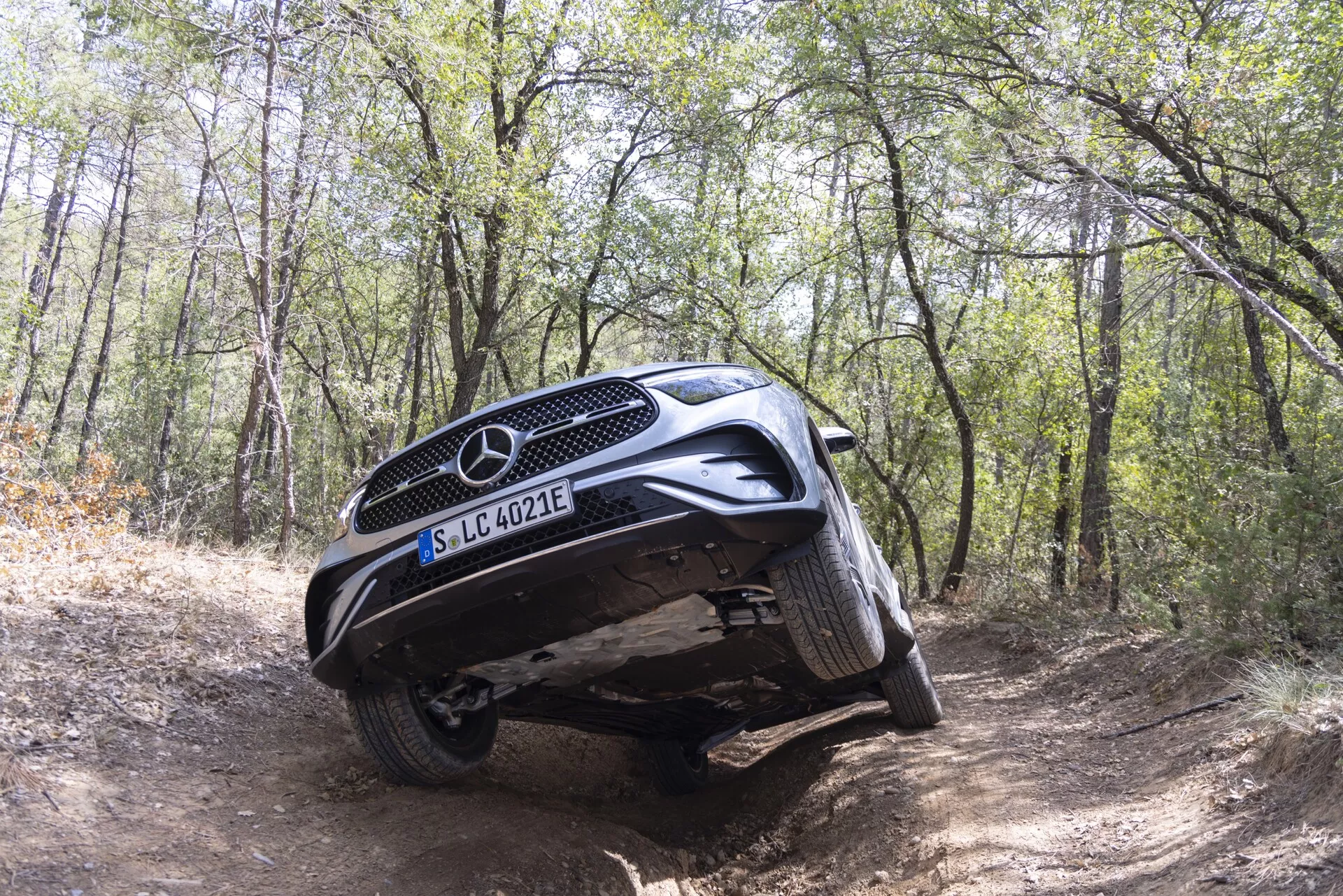Mercedes-Benz GLC 254 z 2022 roku uchwycony w środowisku terenowym w lesie, pokazując swoje zdolności w trudnym terenie. Niski, lekko przedni kąt uwydatnia przedni grill, płytę ochronną i artykulację zawieszenia z uniesionymi lewymi kołami nad garbem i prawymi kołami w rowku. To podkreśla prześwit i terenowe cechy ochronne pojazdu. Emblemat EQ sugeruje technologię elektryczną lub hybrydową. Naturalne światło dzienne, brak sztucznego oświetlenia.