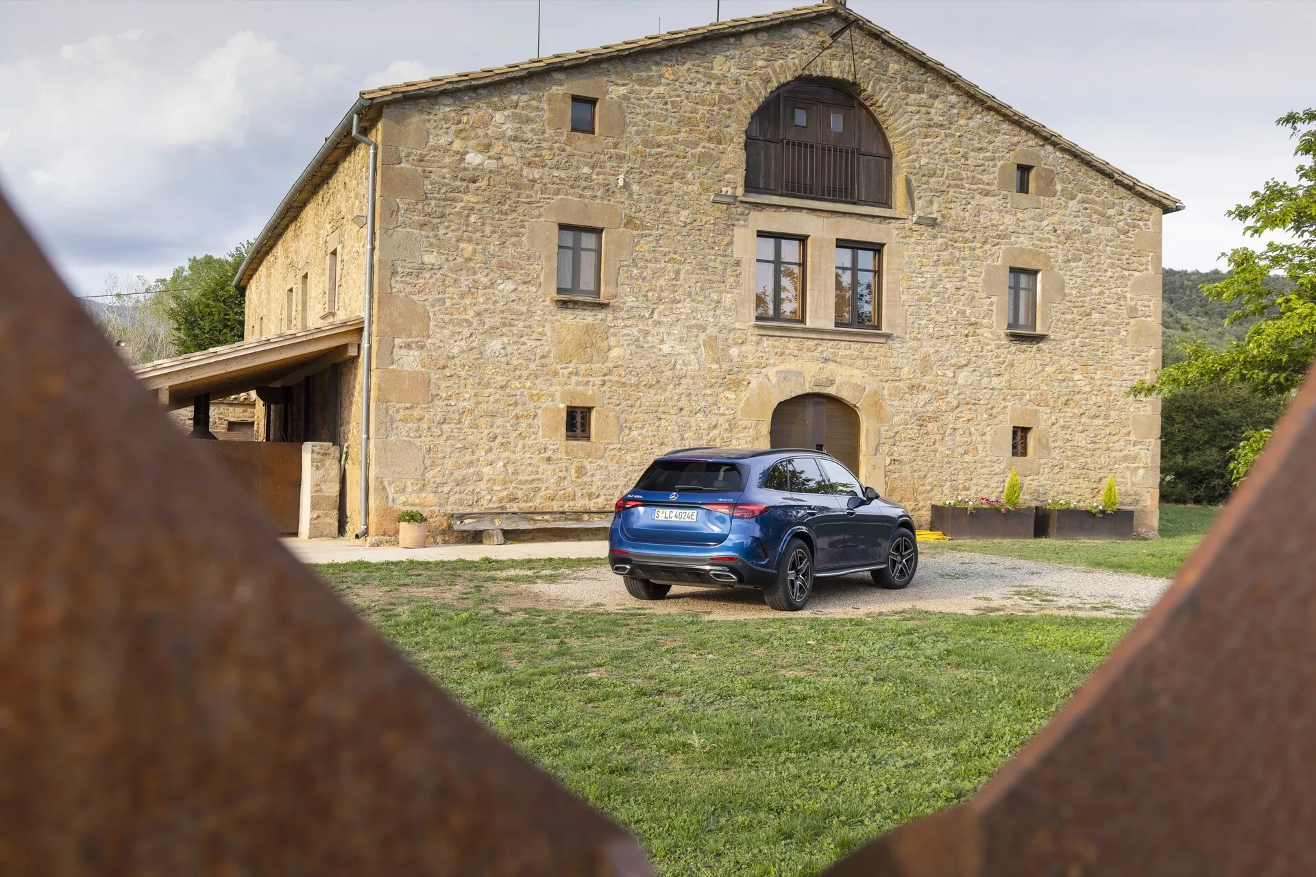 Mercedes GLC 2022 (254) zaparkowany na trawiastym terenie z tradycyjnym kamiennym domem w tle, pokazujący tylny i częściowy boczny profil z tylnymi światłami LED, spojlerem dachowym, relingami dachowymi, tylnymi czujnikami parkowania, felgami aluminiowymi i zardzewiałymi elementami żelaznymi na pierwszym planie.