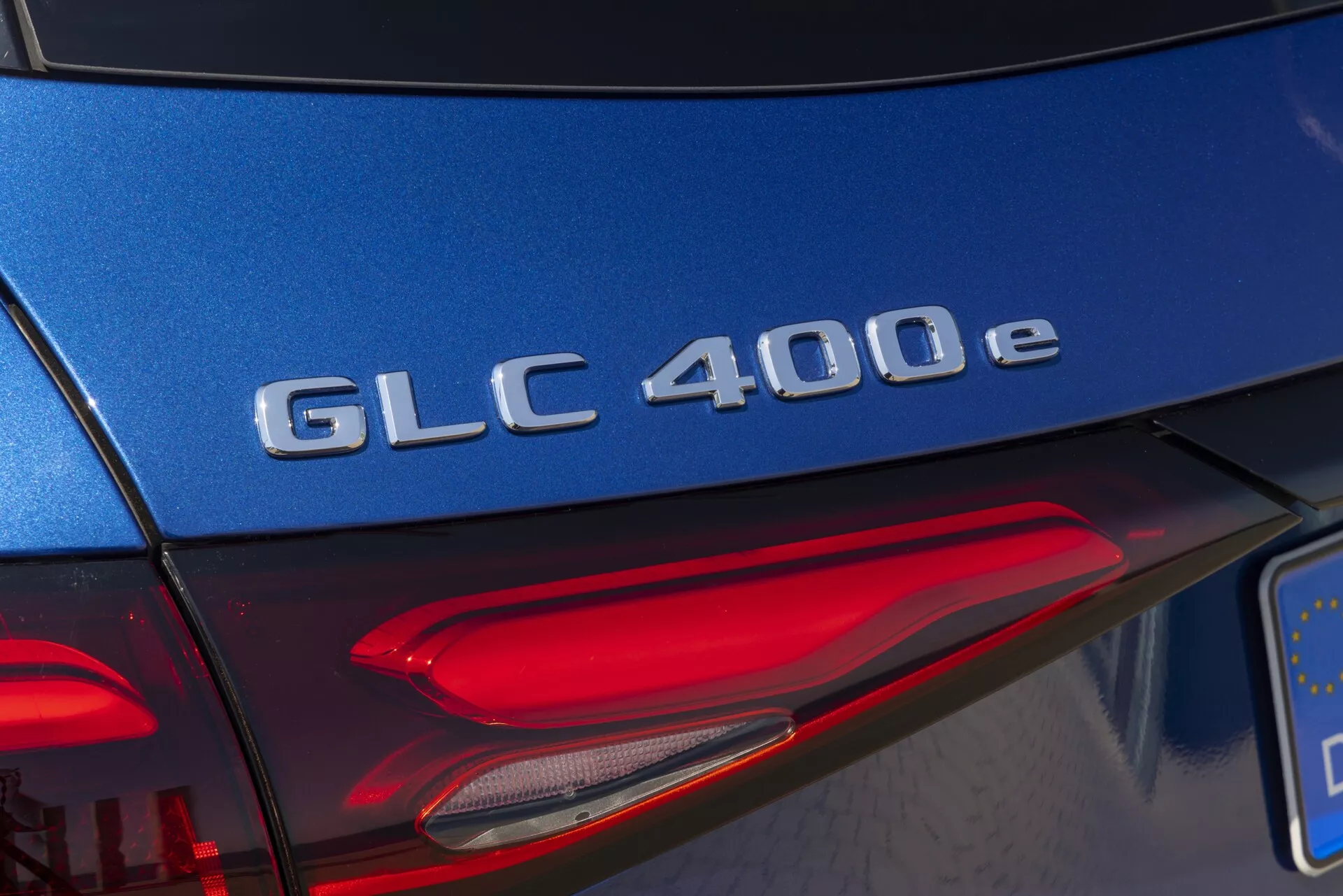 Szczegół tylnego fragmentu modelu Mercedes-Benz GLC 400e 2022, ukazujący zbliżenie oznaczenia modelu w chromie na niebieskich drzwiach bagażnika z ujęciem diagonalnym, ukazujące część lewej tylnej lampy z elementami LED w świetle dziennym.
