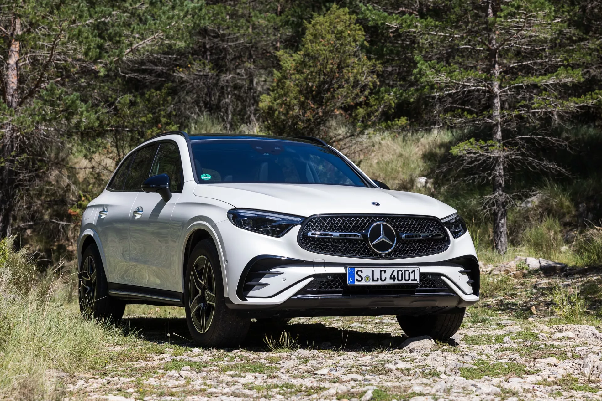 Widok z przodu w trzech czwartych na Mercedes-Benz GLC 2022, podkreślający jego wydajną kratkę, eleganckie reflektory LED, duże wieloszprychowe czarne felgi aluminiowe i aerodynamiczne białe nadwozie. Pojazd zaparkowany na skalistym terenie z drzewami w tle, podkreślając jego luksusowy wygląd i możliwości SUV.