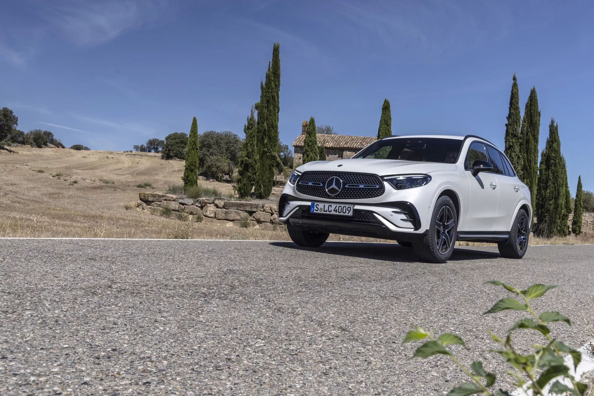 Zdjęcie niskiego kątu białego SUV-a Mercedes GLC 2022 zaparkowanego na drodze z naturalnym krajobrazem w tle, z przejrzystym niebem i drzewami cyprysowymi. Przednia ćwierć pojazdu jest uchwycona od strony kierowcy, pokazując eleganckie reflektory LED, wyraźny przedni grill z dużym emblematem, sportowe felgi i profilowaną maskę. Delikatnie opadająca linia dachu i dobrze zdefiniowana linia ramienia podkreślają projekt łączący aerodynamikę i solidność. Światło słoneczne tworzy cienie i refleksy, podkreślając luksusową i nowoczesną estetykę samochodu.