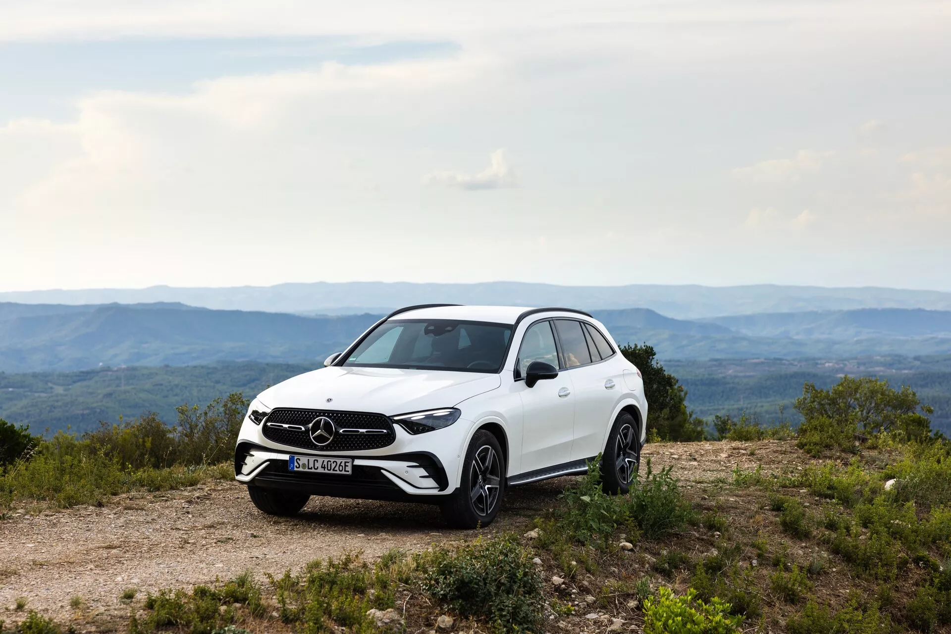 Profil boczny białego Mercedesa GLC 2022 na nierównym szlaku, podkreślający postawę, prześwit i linie projektowe SUV-a. Widoczne funkcje obejmują przednią kratkę i emblemat marki, reflektory LED, obręcze aluminiowe, lusterka boczne z wbudowanymi kierunkowskazami, aerodynamiczne linie nadwozia, błyszczące wykończenie lakieru i wzór bieżnika. Naturalne, dzienne oświetlenie podkreśla estetyczne i funkcjonalne cechy pojazdu.