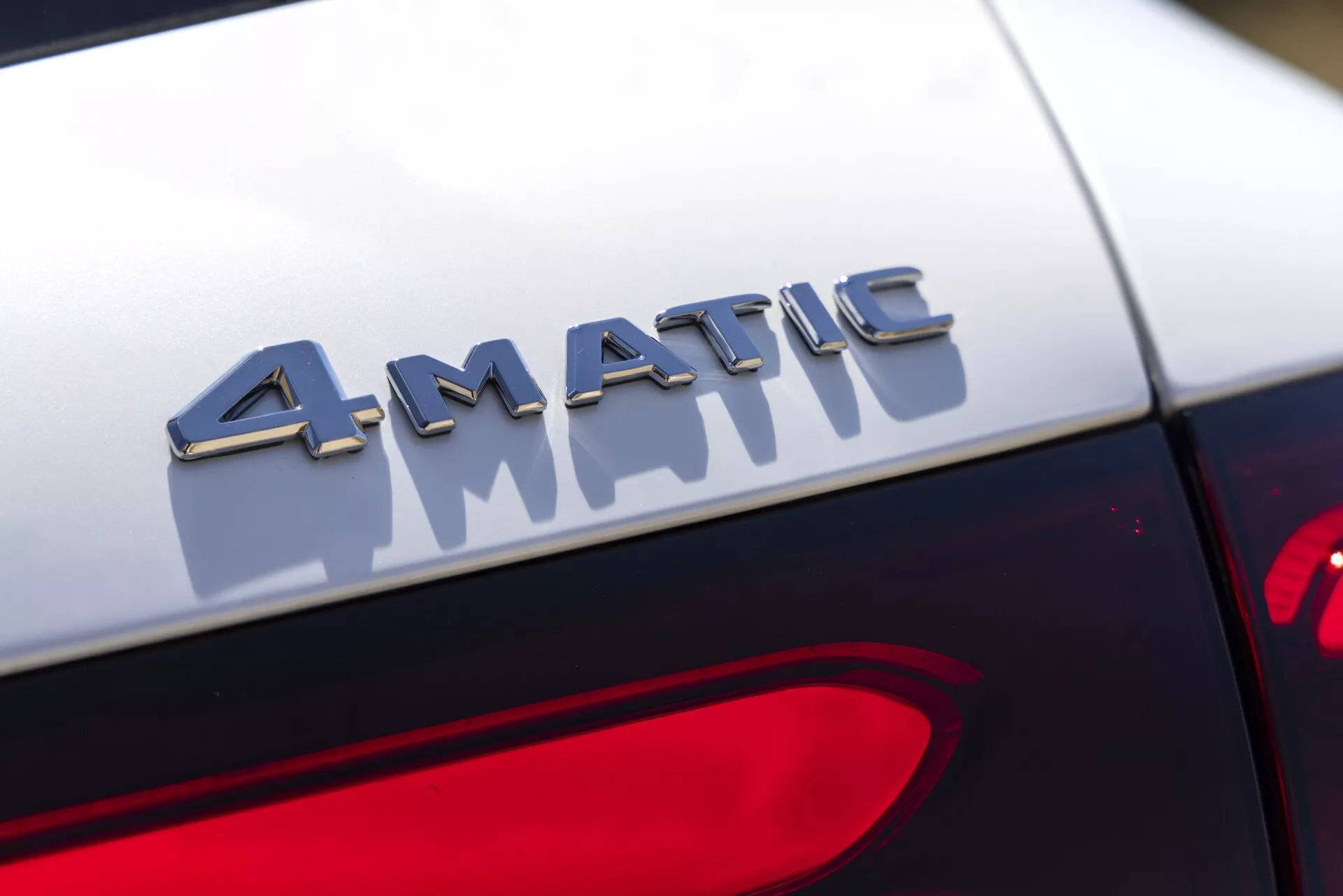 Zbliżenie na detal odznaki 4MATIC na tylnej części samochodu Mercedes GLC 2022. Fotografia uchwytuje odznakę w ostrym skupieniu z metalowymi literami odbijającymi światło, zamontowaną tuż nad światłem tylnym. Tło jest rozmyte, podkreślając trójwymiarowy efekt odznaki i kontrast z ciemnym lakierem samochodu. Projekt tylnego światła częściowo widoczny w prawym dolnym rogu.