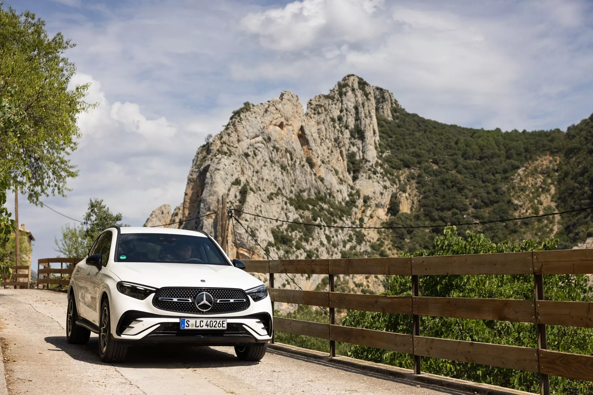 Biały Mercedes-Benz GLC 2022 zaparkowany na malowniczej drodze, widok z przedniego prawego rogu, pokazujący grille, reflektory i część prawej strony, w tym przednie drzwi pasażera i koło. Tablica rejestracyjna 'SLC 400 e' wskazująca na możliwy pojazd hybrydowy typu plug-in lub elektryczny. Uchwycony w ciągu dnia, biały lakier i błyszcząca powierzchnia samochodu odbijają naturalne światło, podkreślając duże logo Mercedes-Benz na grillu. Skaliste góry i drewniana barierka w tle sugerują wiejską lub górską drogę, wskazując na luksusowy wypoczynek lub odkrywanie na świeżym powietrzu.