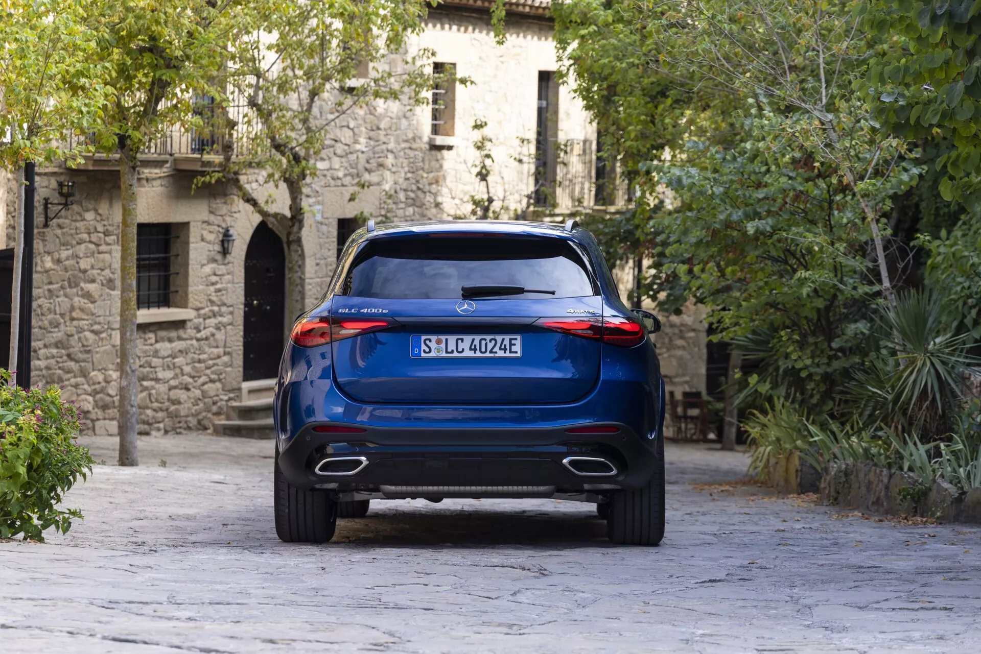 Widok z tyłu niebieskiego SUV-a Mercedes GLC 2022 z lekko niskiej perspektywy, pokazujący jego wysoką wysokość, podwójne wyloty wydechowe i eleganckie tylne światła. Pojazd umieszczony centralnie stoi na tle europejskiej architektury i brukowanej nawierzchni, podkreślając swoją nowoczesność i możliwy napęd elektryczny.