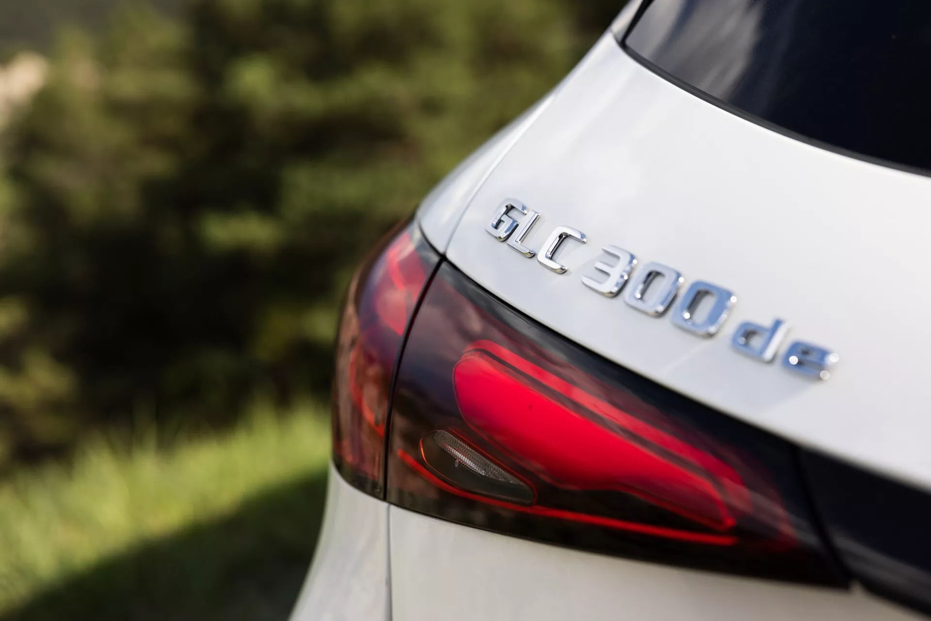 Zbliżenie tylnej części Mercedesa GLC 300de 2022, z naciskiem na plakietkę modelu i część tylnego światła LED. Zdjęcie, wykonane pod nieco podwyższonym kątem bocznym, podkreśla plakietkę GLC 300de i gładki kontur nowoczesnego designu tylnego światła. Metaliczna farba subtelnie odbija światło, sugerując dobrze utrzymaną powierzchnię, choć dokładny kolor wydaje się jasny, prawdopodobnie biały lub srebrny. Płytka głębia ostrości zachowuje ostrość plakietki, jednocześnie rozmywając tło i obszary peryferyjne, podkreślając technologiczne i projektowe detale tego hybrydowego pojazdu z napędem dieslowo-elektrycznym.
