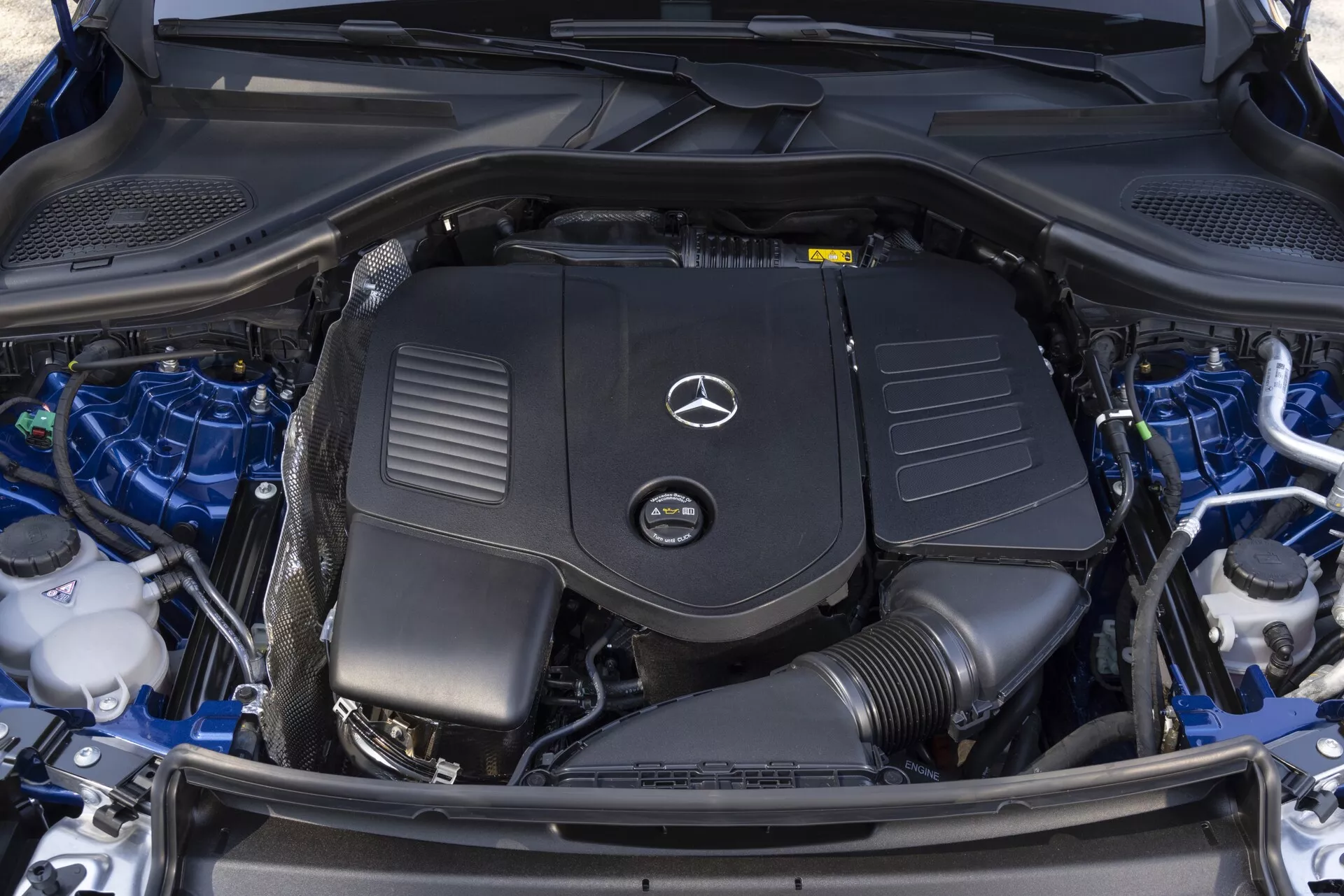 Zbliżenie widoku z góry na czysty i dobrze utrzymany przedział silnika Mercedesa GLC 2022, prezentując pokrywę silnika z wyraźnym logo Mercedes-Benz, akumulator, zbiorniki płynów, elementy systemu chłodzenia i starannie zorganizowaną wiązkę przewodów.
