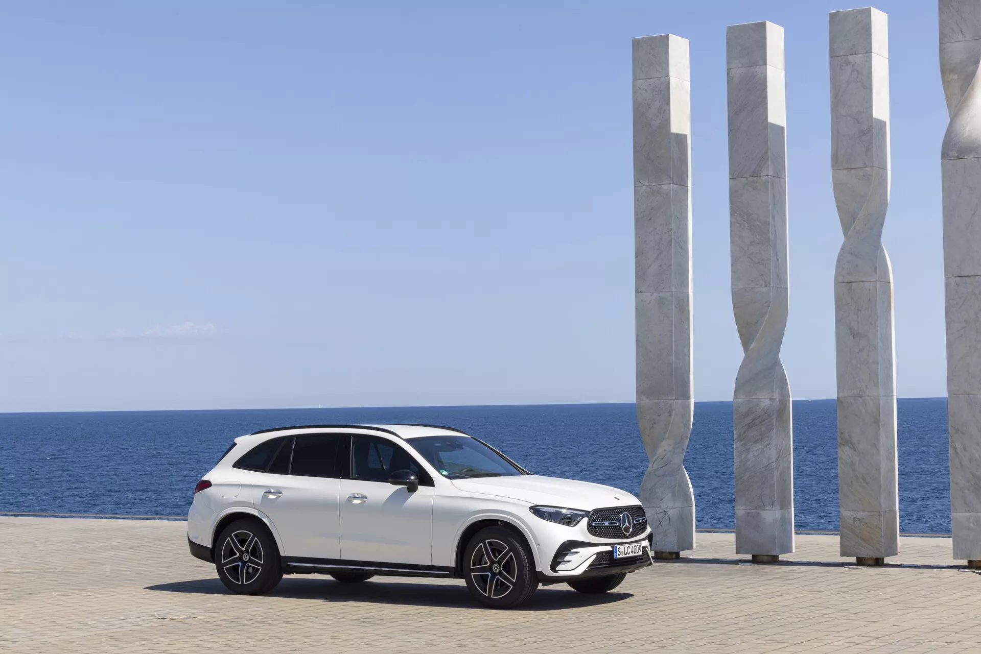 Nowoczesny biały luksusowy Mercedes GLC 2022 zaparkowany z widokiem na malownicze wybrzeże; profil boczny ukazujący elegancki design z podwyższonym prześwitem, dużymi aluminiowymi kołami, zaawansowanymi reflektorami LED, aerodynamicznymi lusterkami z zintegrowanymi kierunkowskazami, opadającą linią dachu w stylu coupé, panoramicznym dachem i przyciemnianymi szybami. Malownicze tło z nowoczesnymi białymi rzeźbami, czystym niebem i spokojnym morzem podkreślają luksusowy charakter samochodu.
