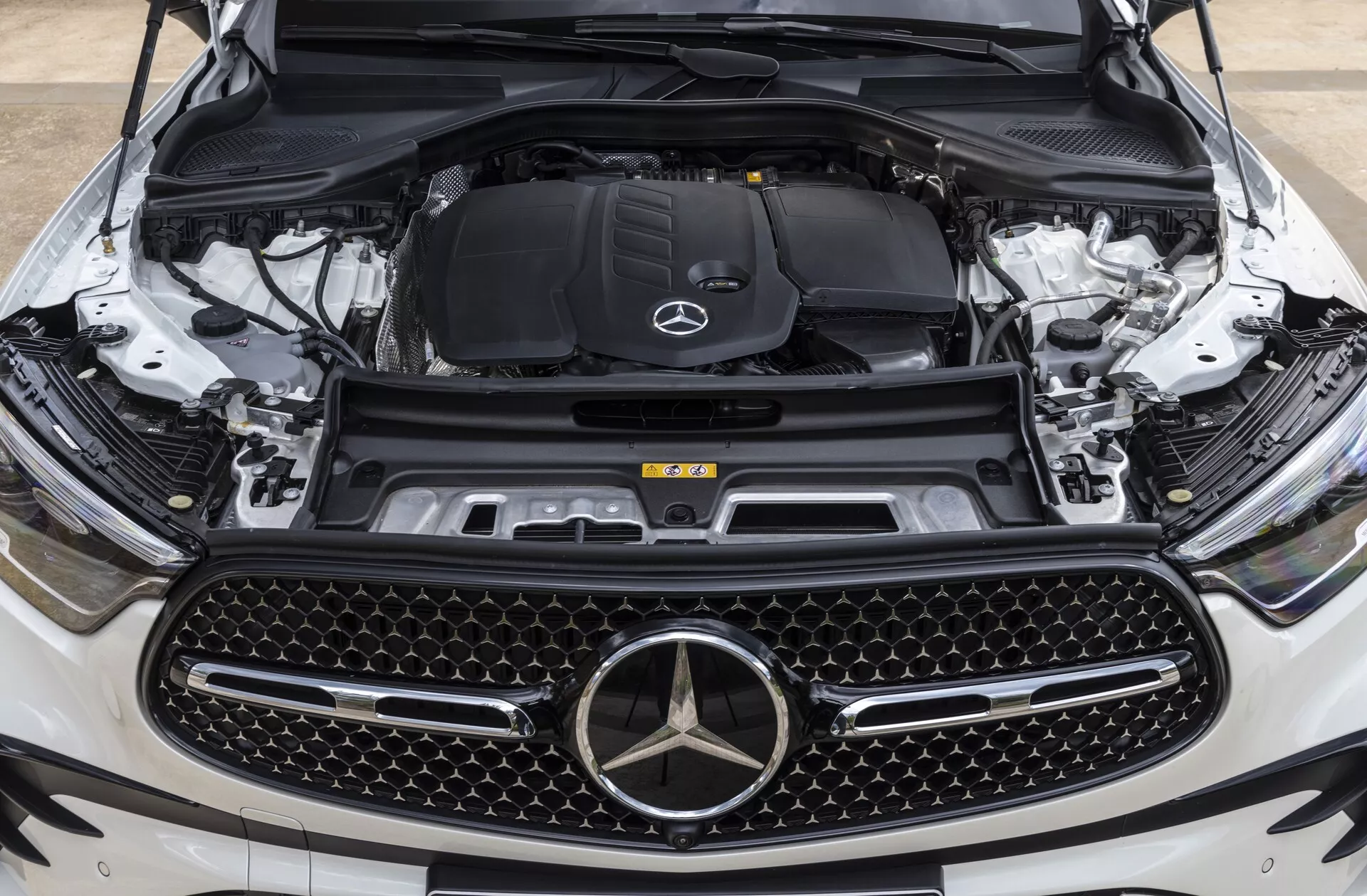 Zbliżony przód Mercedes-Benz GLC 2022 (254) z otwartą maską, pokazujący komorę silnika z perspektywy na wysokości oczu skoncentrowanej na emblemacie marki i grillu, podkreślający duży grill w kształcie diamentu, emblemat gwiazdy Mercedes-Benz, komorę silnika z logo i widoczne komponenty jak blok silnika, układ dolotowy, zbiornik płynu chłodzącego, wieżę zawieszenia i przednią kamerę, wskazującą zaawansowane funkcje techniczne.