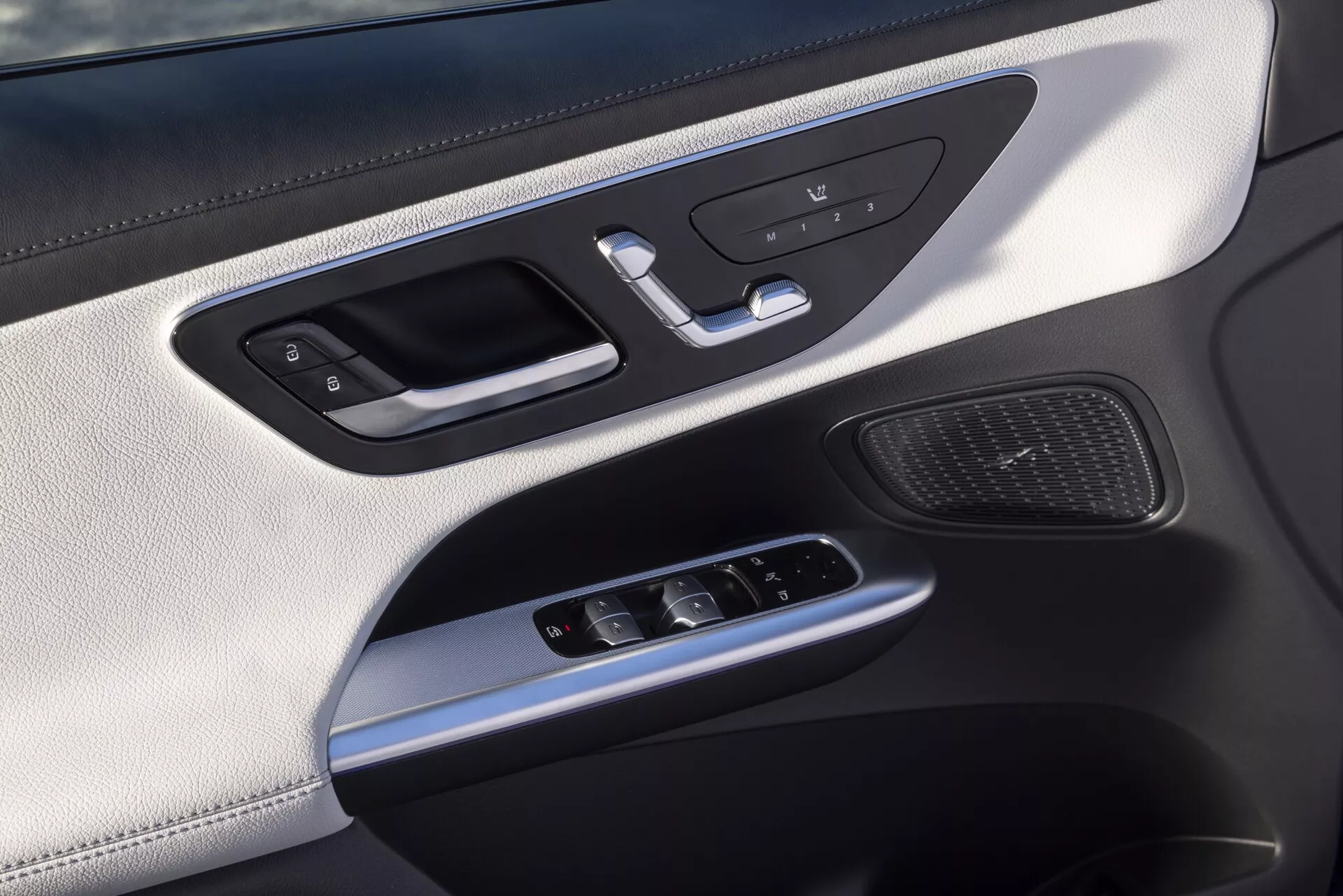 Zbliżenie na panel drzwi wewnętrznych Mercedesa GLC 2022 254, skierowane na stronę kierowcy lub pasażera, zawierające srebrną klamkę z zintegrowanym mechanizmem blokady, przyciski pamięci siedzenia, przełączniki elektrycznych szyb, joystick do regulacji lusterek bocznych, kratkę głośnika, luksusowe materiały w tym górna część podobna do skóry, srebrne akcenty i oświetlenie ambientowe.