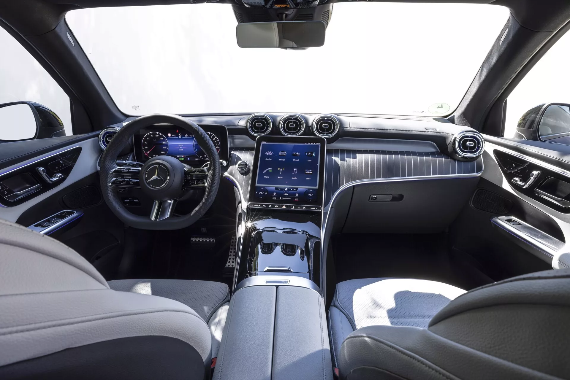 Wnętrze Mercedesa GLC 2022, widok z tylnego środkowego siedzenia. Luksusowa kabina z dwukolorową skórzaną tapicerką w kolorze kremowym i ciemnymi akcentami. Deska rozdzielcza z eleganckim wykończeniem drewnianym oraz dużym pionowym systemem infotainment z ekranem dotykowym, wlotami powietrza i przyciskami sterowania. Widać również wielofunkcyjną kierownicę pokrytą skórą z metalicznymi akcentami oraz cyfrowy zestaw wskaźników.