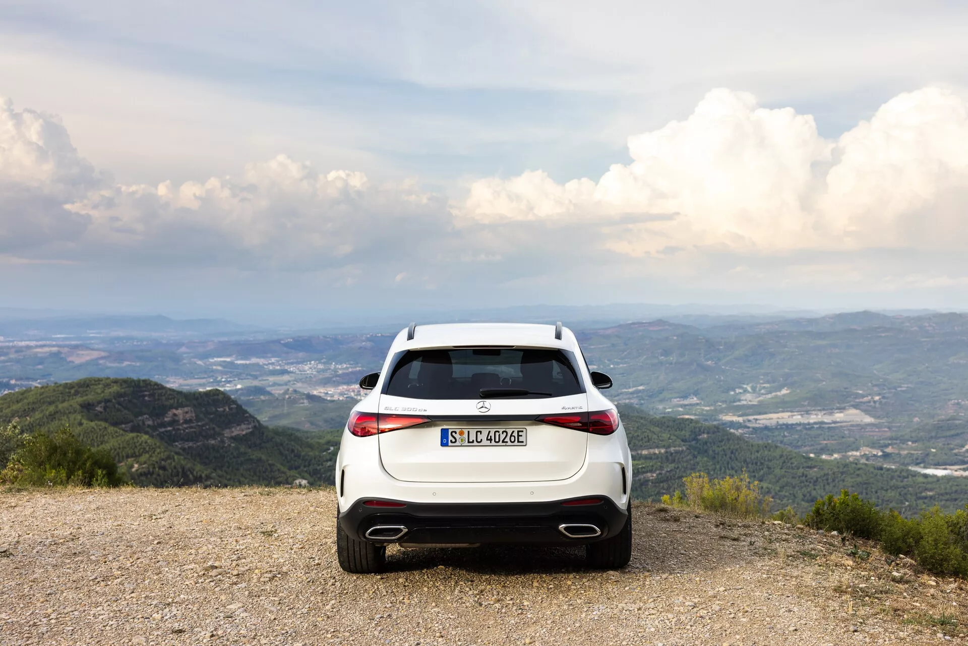 Widok od tyłu białego SUV-a hybrydowego/elektrycznego Mercedes-Benz GLC 2022 (EQ) zaparkowanego na żwirowej powierzchni z malowniczym tłem gór i drzew, pod częściowo zachmurzonym niebem. Zdjęcie podkreśla poziome tylne światła LED, podwójne wydechy, tylny dyfuzor i oznaczenie EQ. Kompozycja sugeruje tematy przygody i ekologicznej podróży.