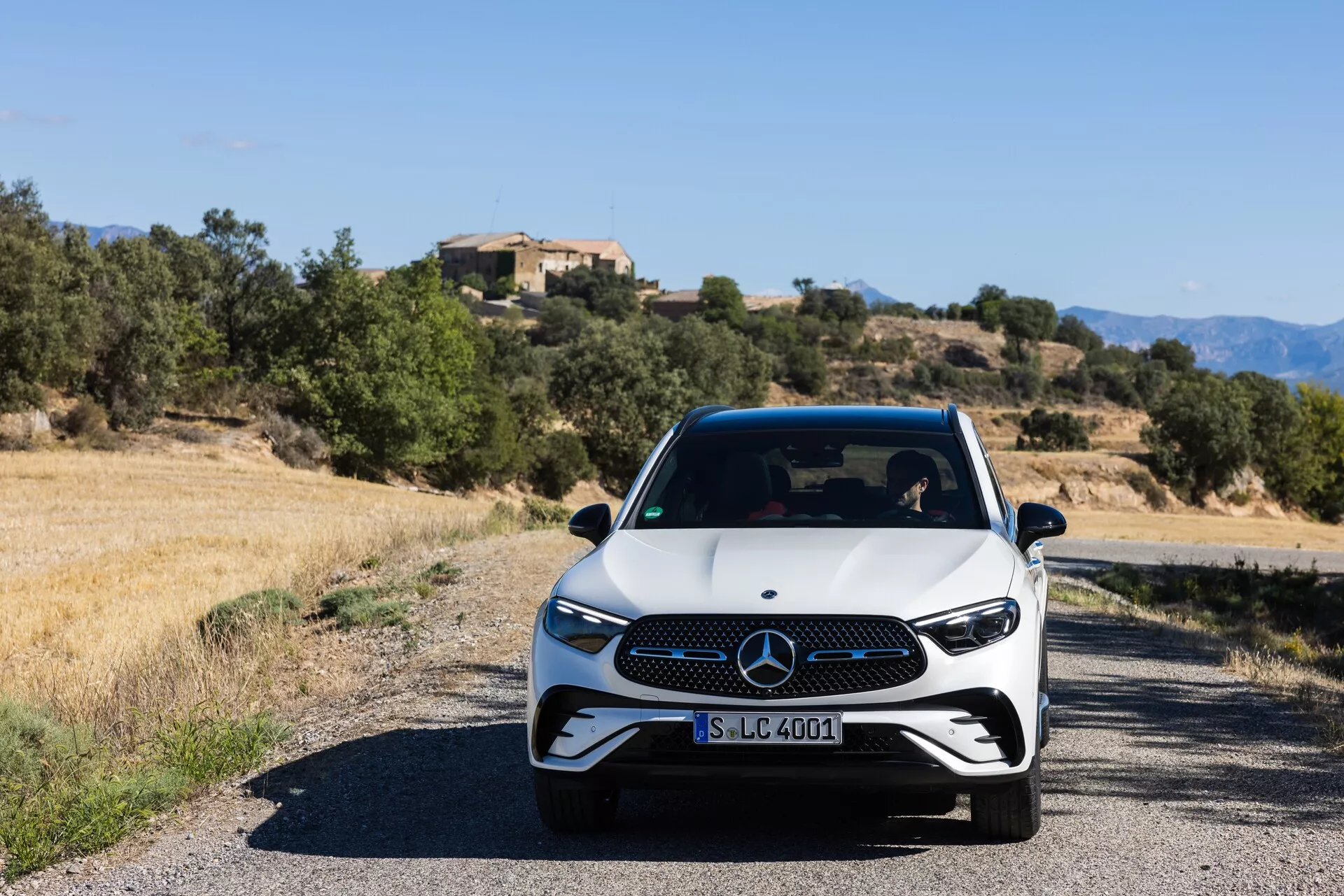 Widok z przodu nowoczesnego Mercedesa-Benz GLC 2022 (254) na wąskiej drodze, z białą bazą i czarnymi detalami, reflektorami LED i charakterystycznym grillem. Zdjęcie zrobione na poziomie oczu w ciągu dnia ukazuje sportowy i premium wykończenie pojazdu z śródziemnomorskim krajobrazem w tle.