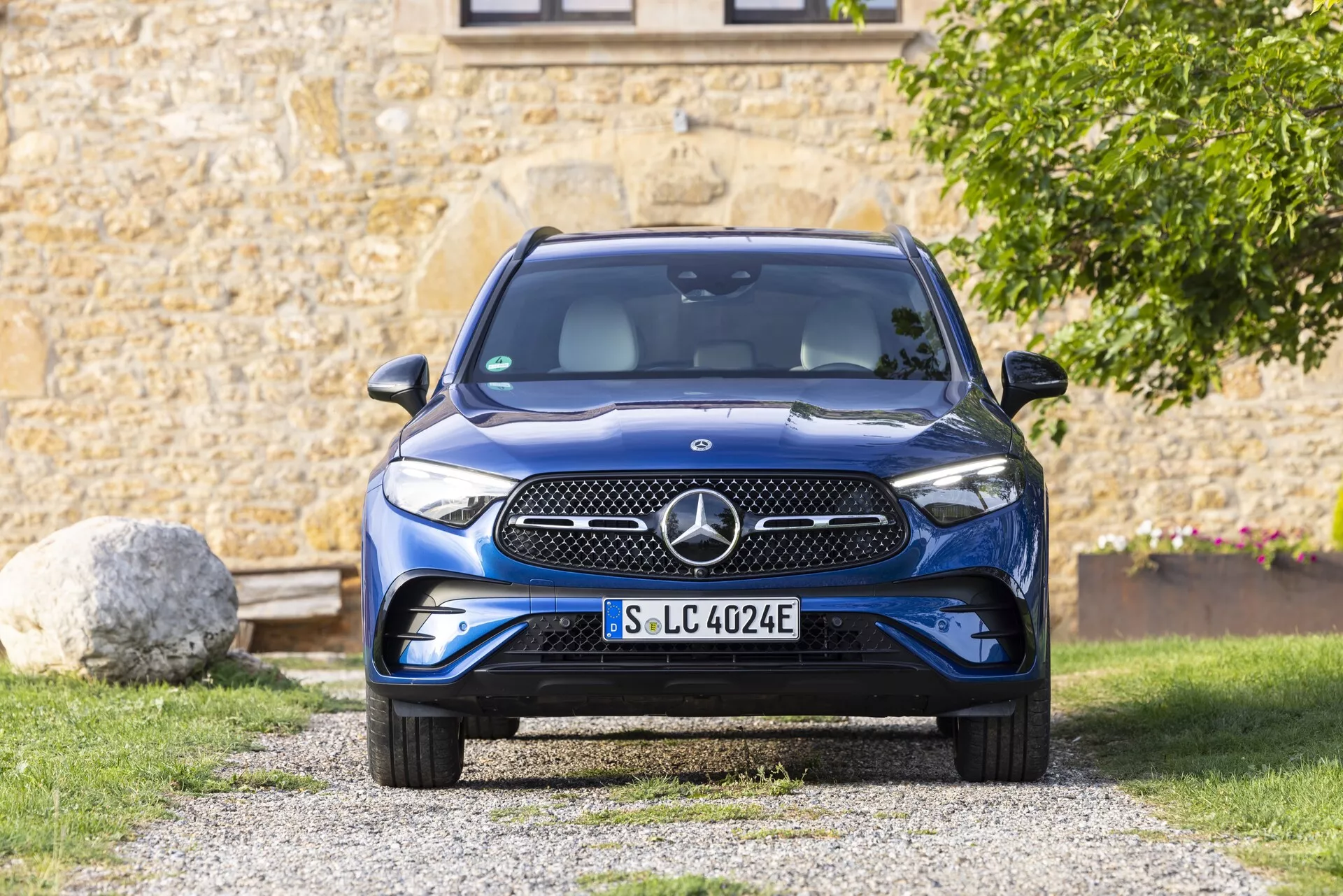 Widok z przodu na Mercedesa GLC 2022 (254) ukazujący wyrazisty przedni grill, zderzak, maskę, reflektory i przednią szybę. Samochód jest umieszczony na podjeździe w podmiejskim otoczeniu z zielenią i częścią budynku w tle. Grill ma gładką powierzchnię z emblematem prawdopodobnie zawierającym czujniki lub kamerę. Eleganckie reflektory wydają się mieć technologię LED. Dolny zderzak zawiera wloty powietrza i czujniki parkowania. Niebieski akcent na tablicy rejestracyjnej wskazuje, że jest to pojazd elektryczny (EV) ze specyficznymi funkcjami EV. Duża przednia szyba zapewnia dobrą widoczność i prawdopodobnie zawiera wycieraczki z czujnikiem deszczu i czujnikiem światła. Lusterka boczne, choć nie są w pełni widoczne, są montowane na drzwiach i mogą mieć monitorowanie martwego pola. Koła mają aerodynamiczny kształt, który poprawia efektywność zasięgu. Obraz jest dobrze oświetlony światłem naturalnym, które podkreśla szczegóły i kolor samochodu.