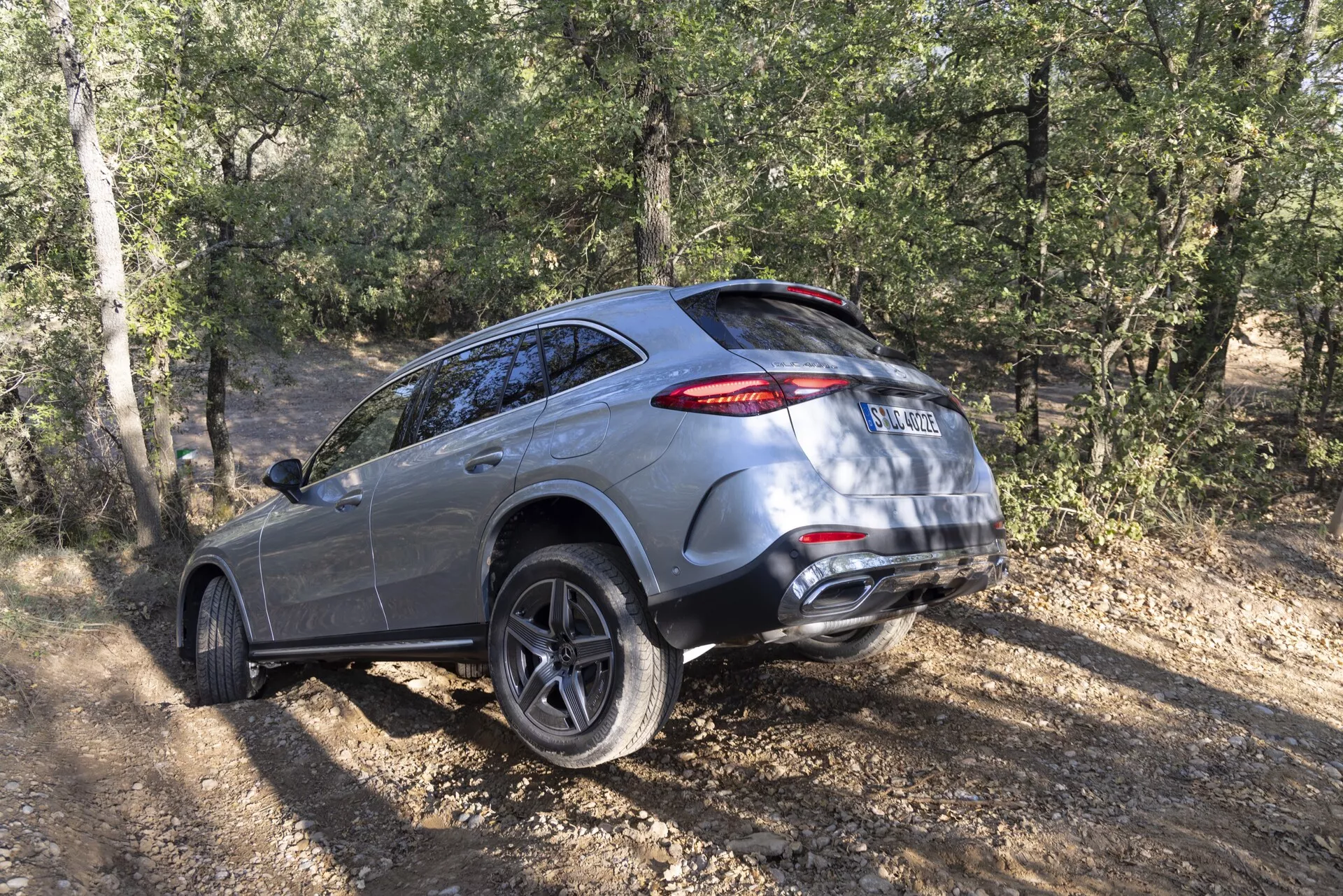 Widok trzy-czwarte tyłu SUV-a Mercedes GLC 2022 na nieutwardzonym terenie, pokazujący jego luksusowe i nowoczesne cechy, takie jak wieloramienne felgi aluminiowe, tylne światła LED, podwójny układ wydechowy i terenowe możliwości z tłem roślinności i drzew.