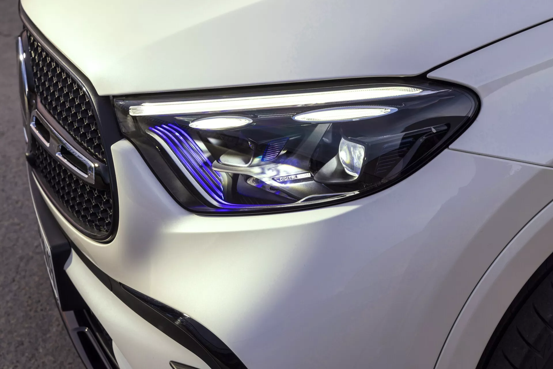 Zbliżenie na przedni lewy róg Mercedes GLC 2022, ukazujące elegancki, nowoczesny design reflektorów z ostrymi, kanciastymi kształtami i zaawansowanymi elementami oświetleniowymi, w tym LED-owe światła do jazdy dziennej i niebieski akcent. Błyszczący biały lakier podkreśla dobrze zdefiniowane linie pojazdu, a widoczna kratka w kształcie plastra miodu odzwierciedla jego sportowy styl.