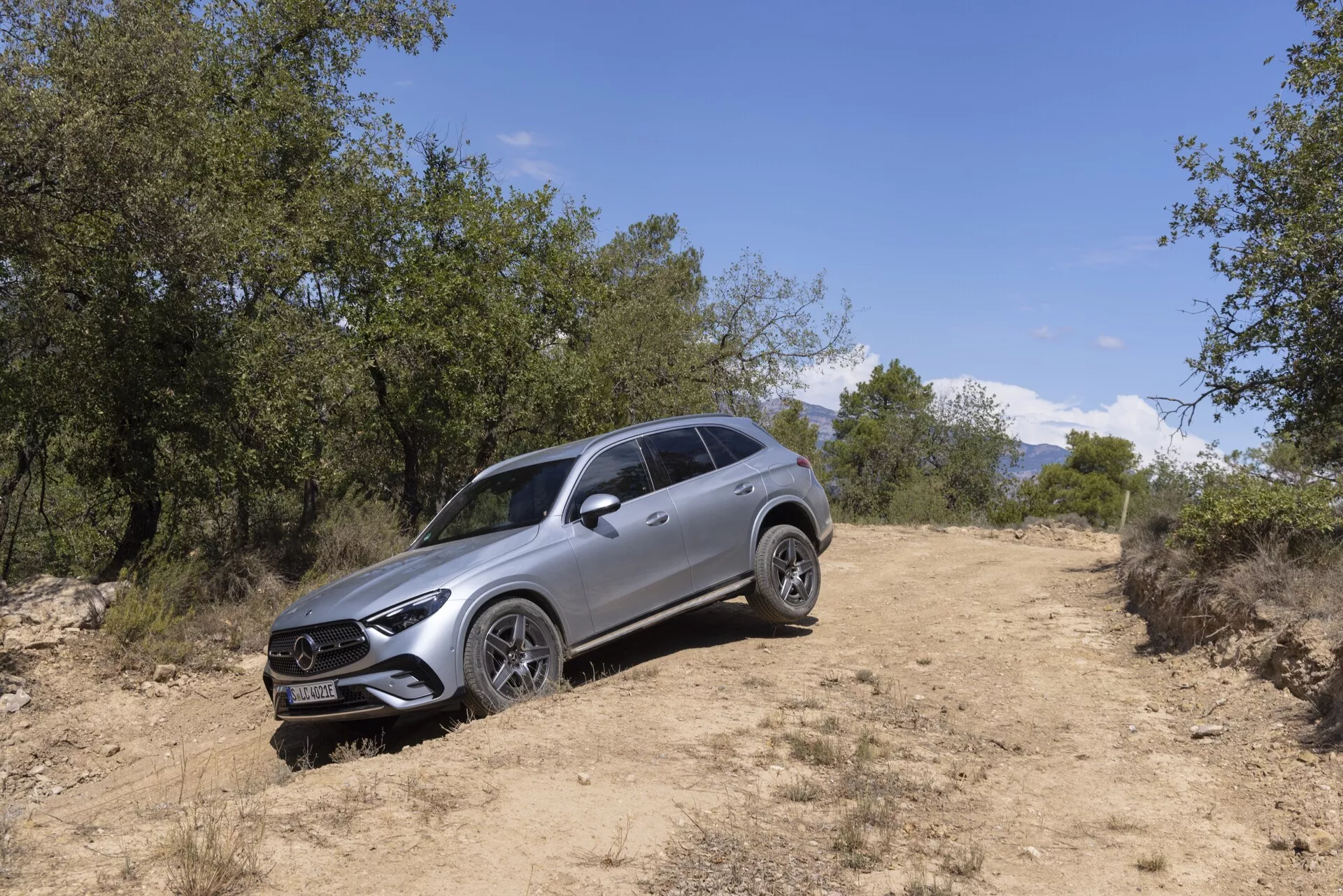 Zdjęcie SUV-a Mercedes GLC 2022 w naturalnym, terenowym otoczeniu uchwycone pod kątem trzy czwarte od przedniej lewej strony. Widoczne cechy obejmują wyrazisty grill z centralnym emblematem, reflektory LED z światłami do jazdy dziennej, wieloramienne obręcze aluminiowe, relingi dachowe, wyrzeźbioną karoserię, lusterka boczne z zintegrowanymi kierunkowskazami oraz podwyższony prześwit wskazujący na możliwości terenowe. Pojazd znajduje się na polnej ścieżce otoczonej trawą i drzewami, z naturalnym światłem słonecznym oświetlającym jego nowoczesny design i luksusowe cechy.